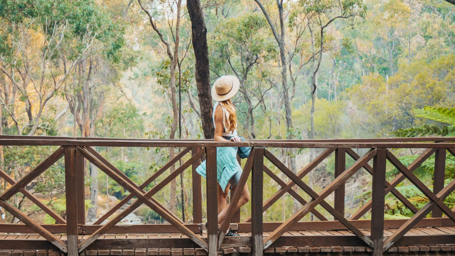 A woman with long hair wearing a wide hat, a white tank top, denim shorts, and a teal quilted jacket draped over her arm, standing on a wooden bridge exploring a forest with green and yellow foliage.