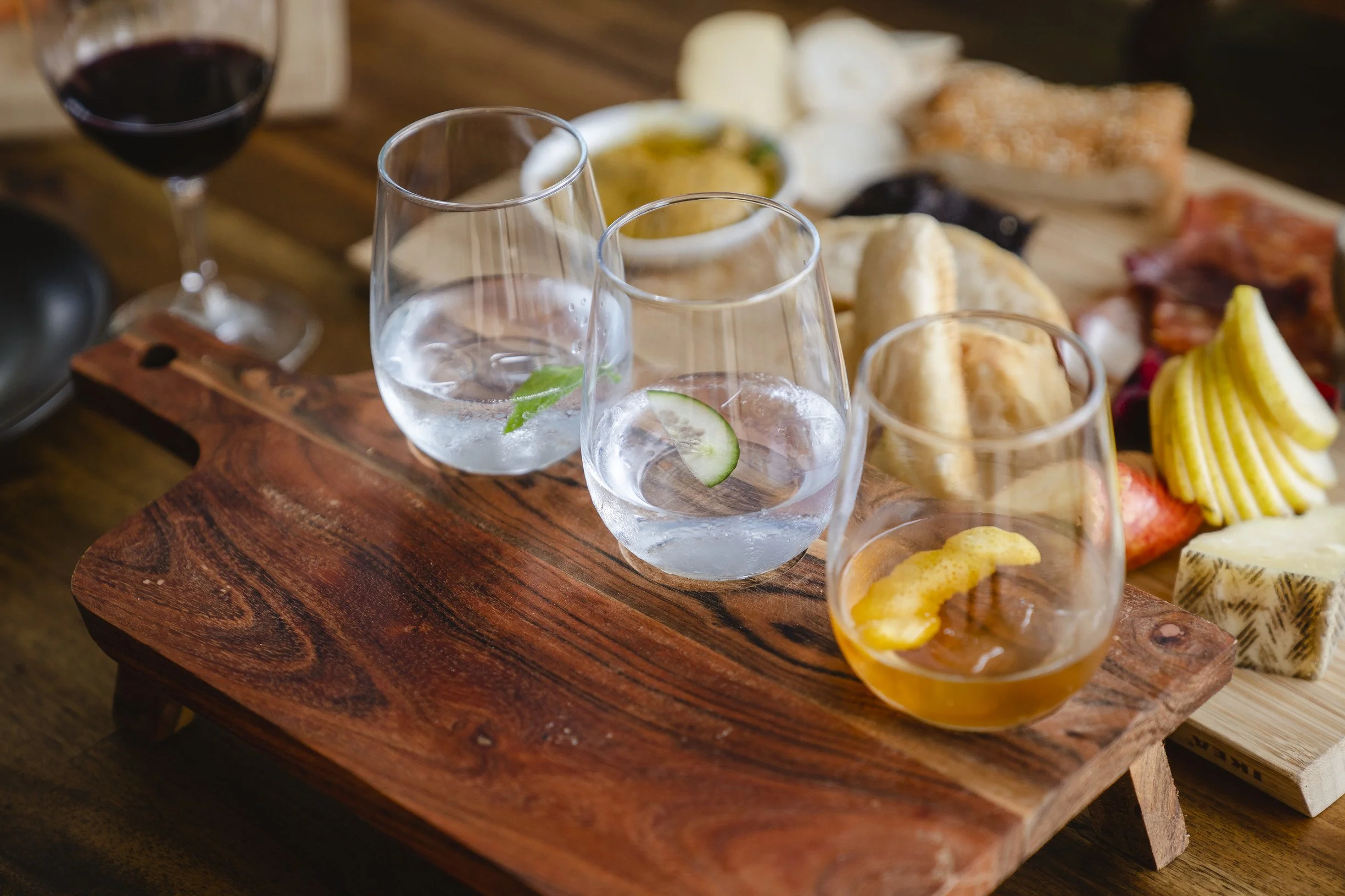 Close-up of a wooden serving board with three glasses of water, some bread rolls, cheese, cold cuts, and a small bowl of green sauce or dip, placed on a wooden table.