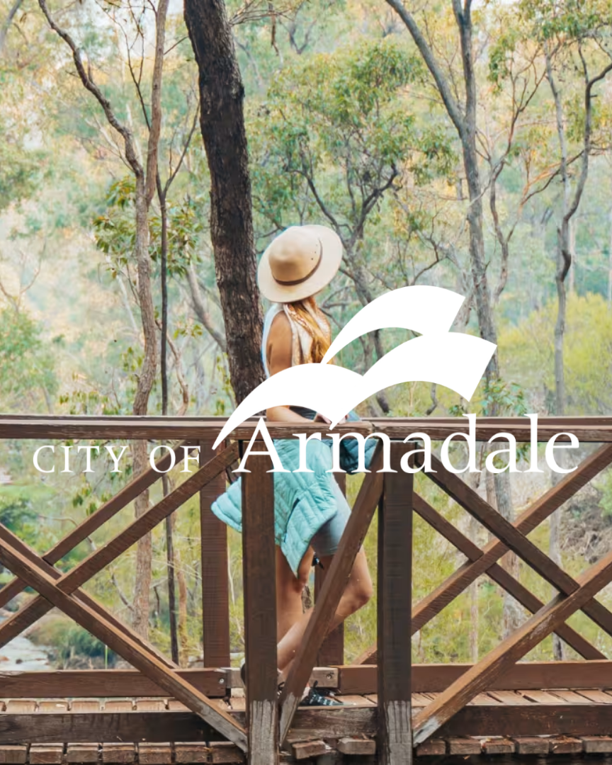 Woman with a hat and towel standing on a wooden bridge in a forested area, with the text "City of Armadale" and a logo overlayed.
