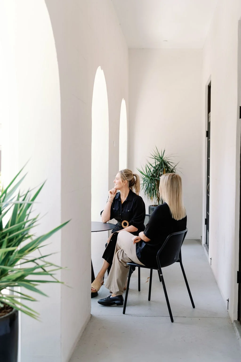 Two women sit on black chairs in a bright, minimalist corridor with large arched windows and potted plants, engaging in conversation.