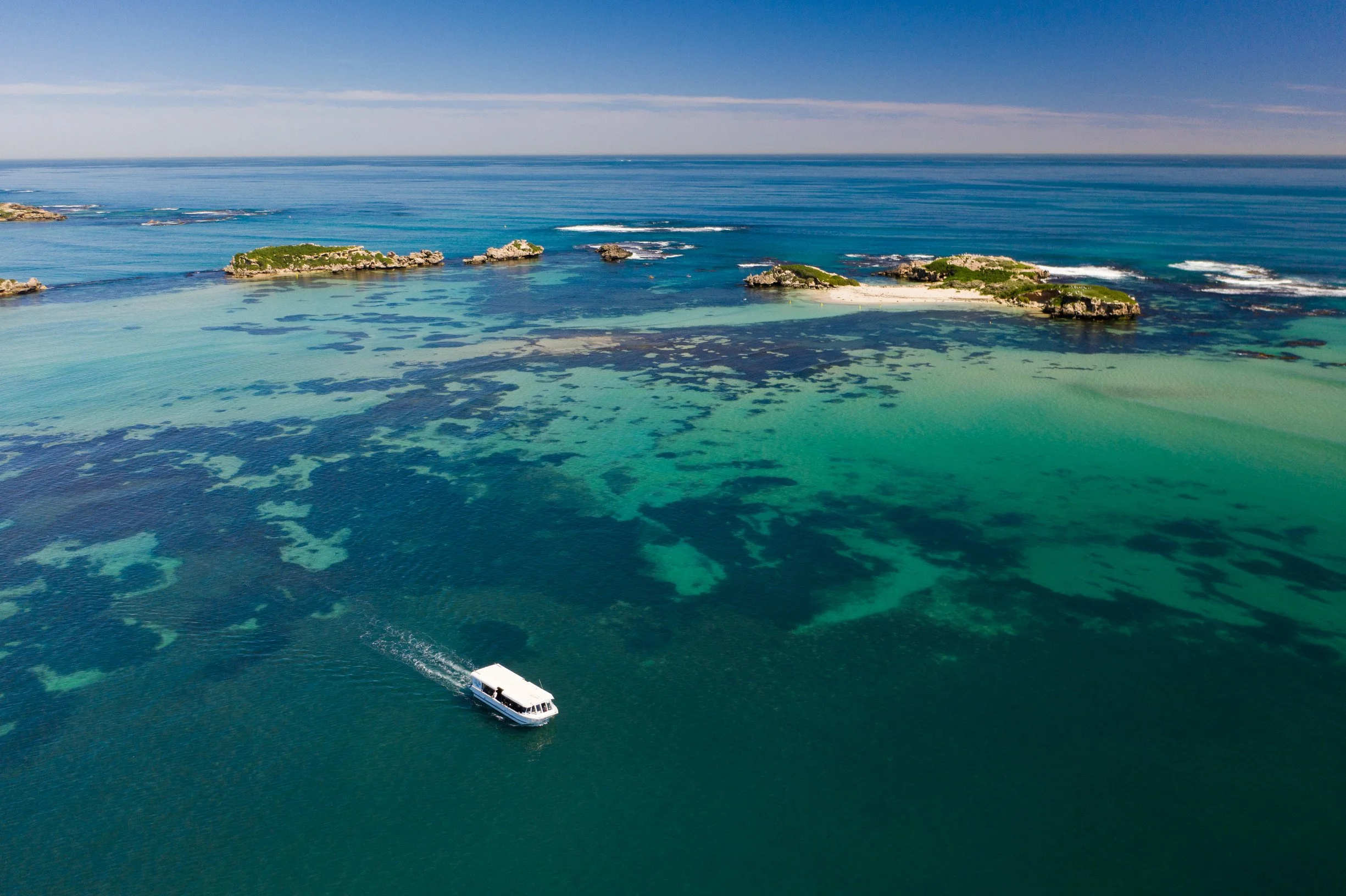 Aerial view of a cruise ship sailing near small islands with lush green vegetation surrounded by turquoise and deep blue ocean water.