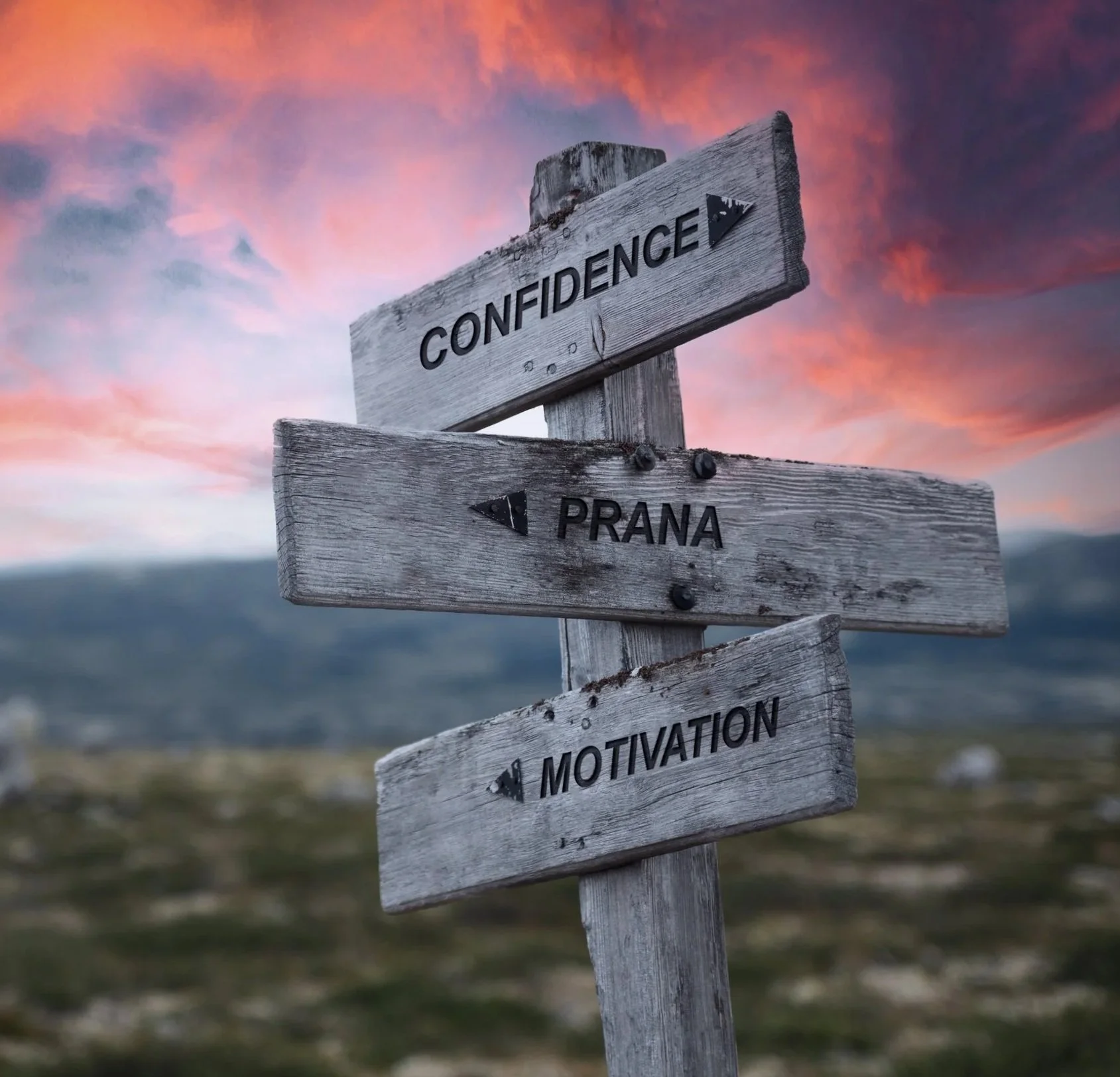 A weathered wooden signpost with three directional signs, reading 'CONFIDENCE,' 'PRANA,' and 'MOTIVATION,' against a scenic backdrop of a colorful sunset sky and distant mountains.