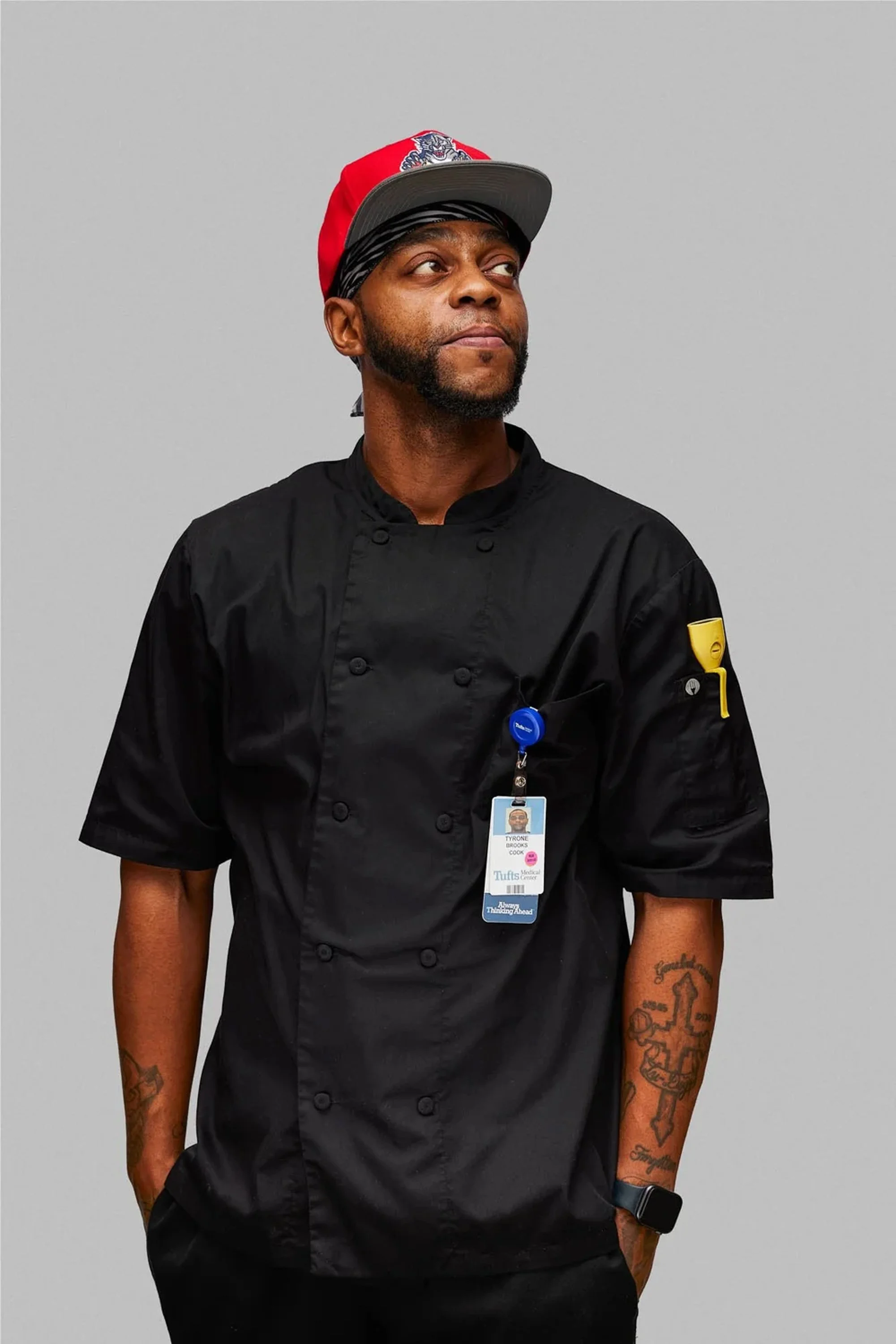 A man dressed as a chef with tattoos on his arms, wearing a black chef's coat, a black smartwatch, and an employee badge hanging from his neck. He is also wearing a red cap with a logo, a black and white bandana, and a black cap underneath. The backg