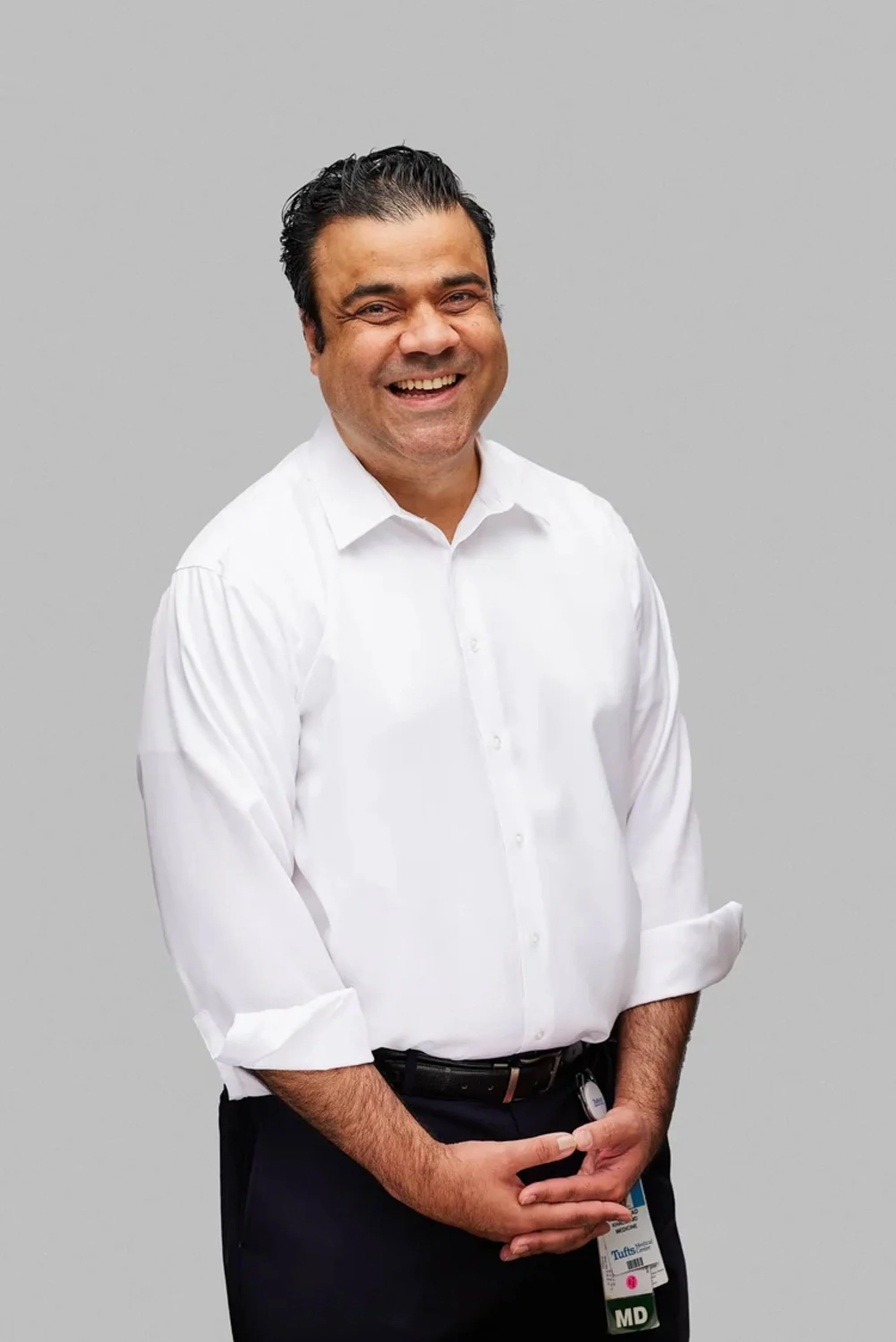 A man in a white shirt and dark pants standing against a plain gray background. He is smiling and holding an ID badge and badge holder in his hands, which are in front of him.