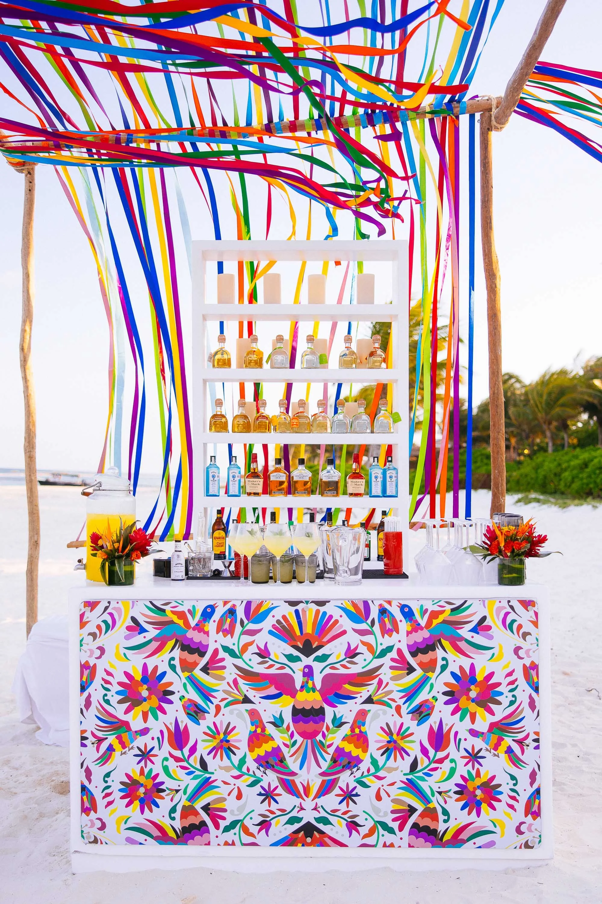Colorful beach bar backdrop with bottles of alcohol, cocktails, flowers, and vibrant decorations on a sandy beach.