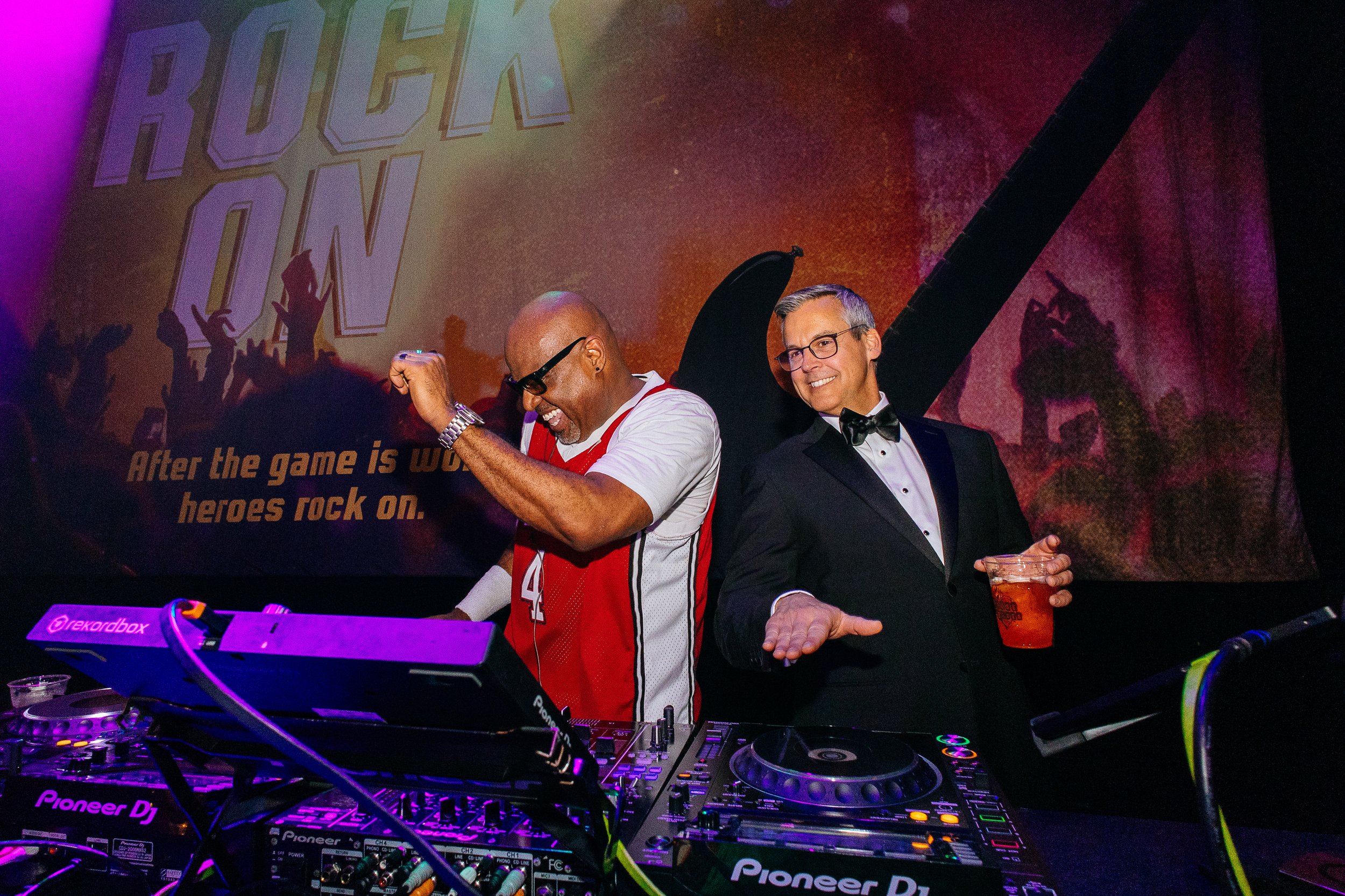 Two men at a party, one dressed as a DJ and the other in a tuxedo, dancing and enjoying drinks with a rock-themed backdrop behind them.