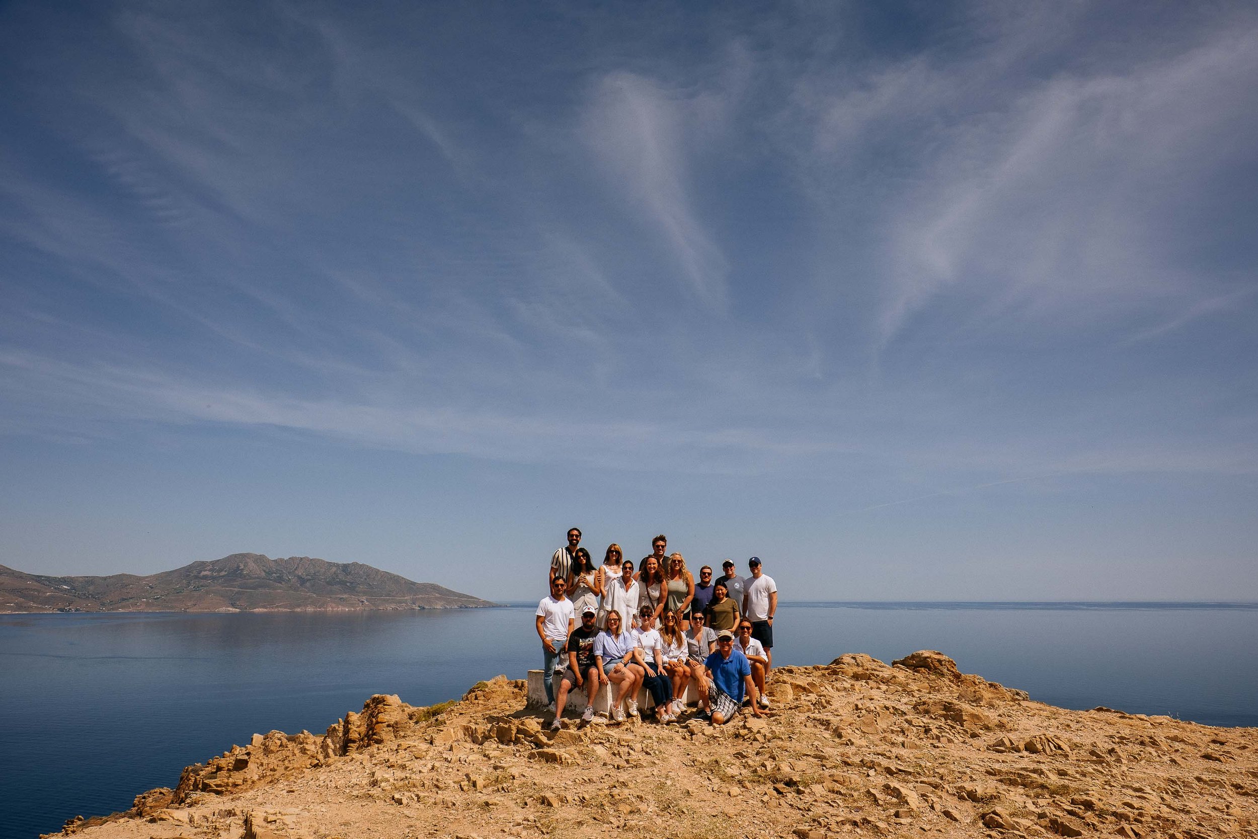 Group of people posing on rocky terrain by the water with mountains in the background and a blue sky with wispy clouds.