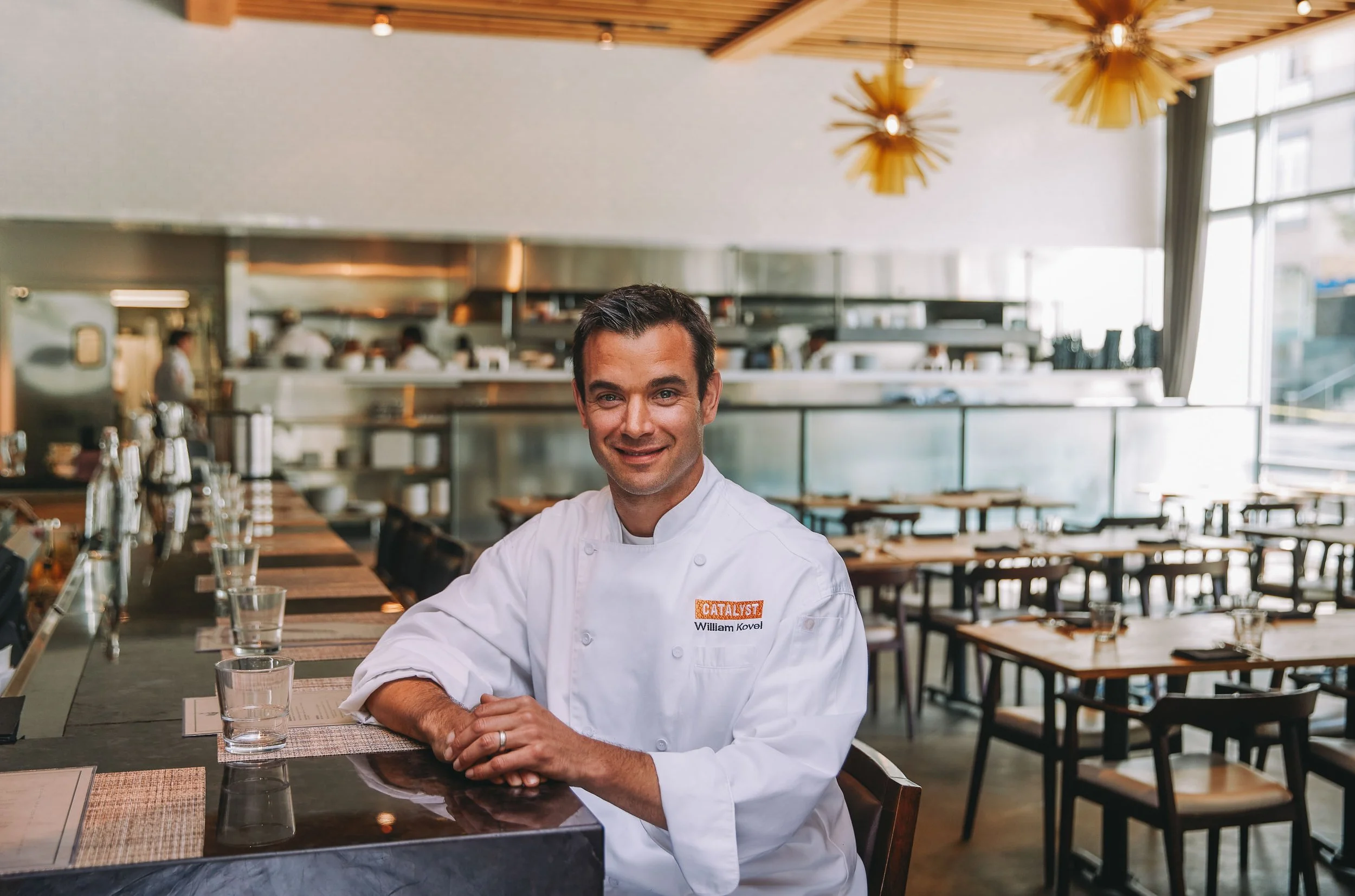 A male chef wearing a white uniform with a name tag that reads William Kovel, sitting at a restaurant table, smiling. The restaurant has large windows, modern decor, and a visible open kitchen in the background.