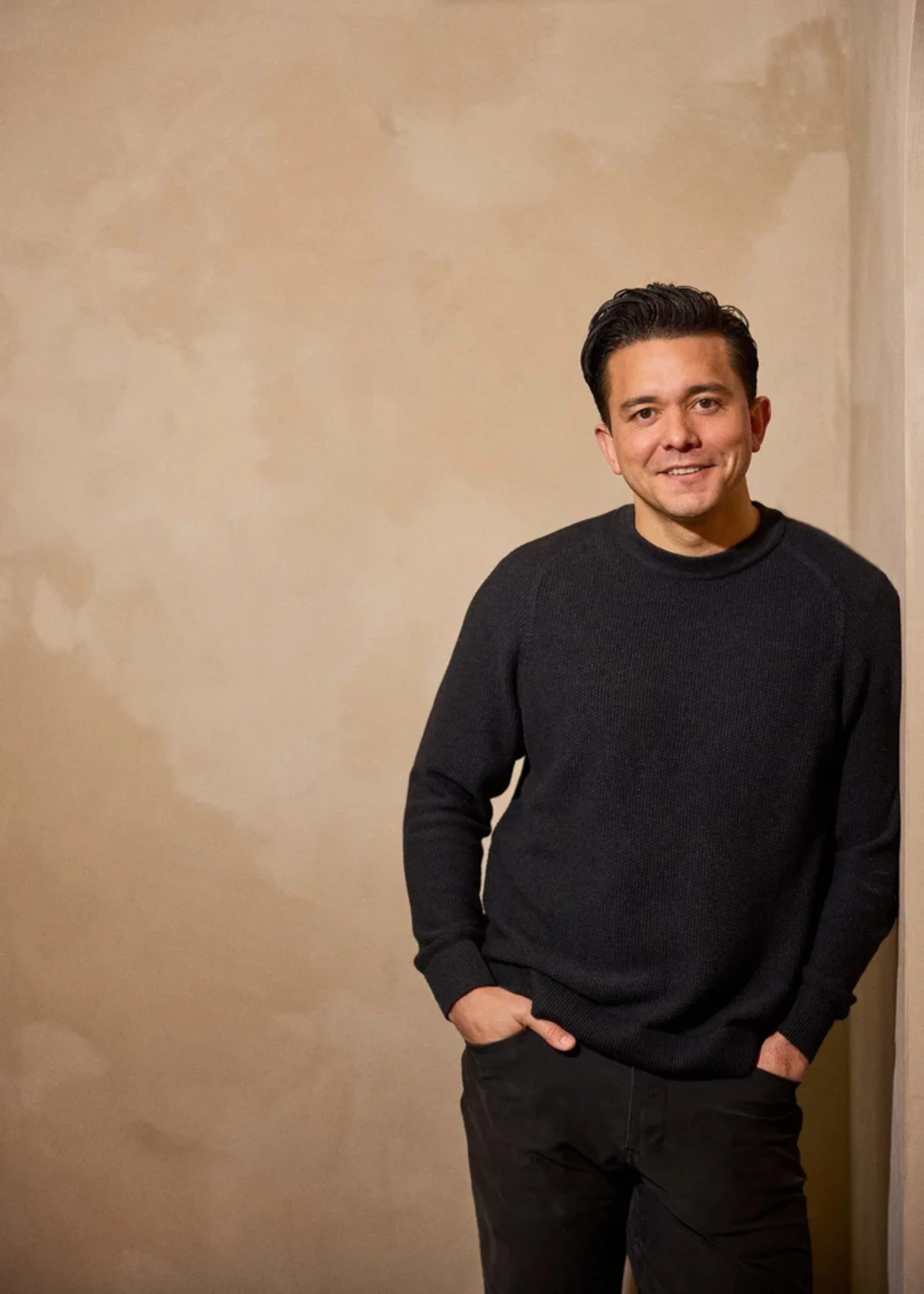 A man with dark hair, wearing a black sweater and pants, standing in front of a plain beige wall, smiling with one hand in his pocket.