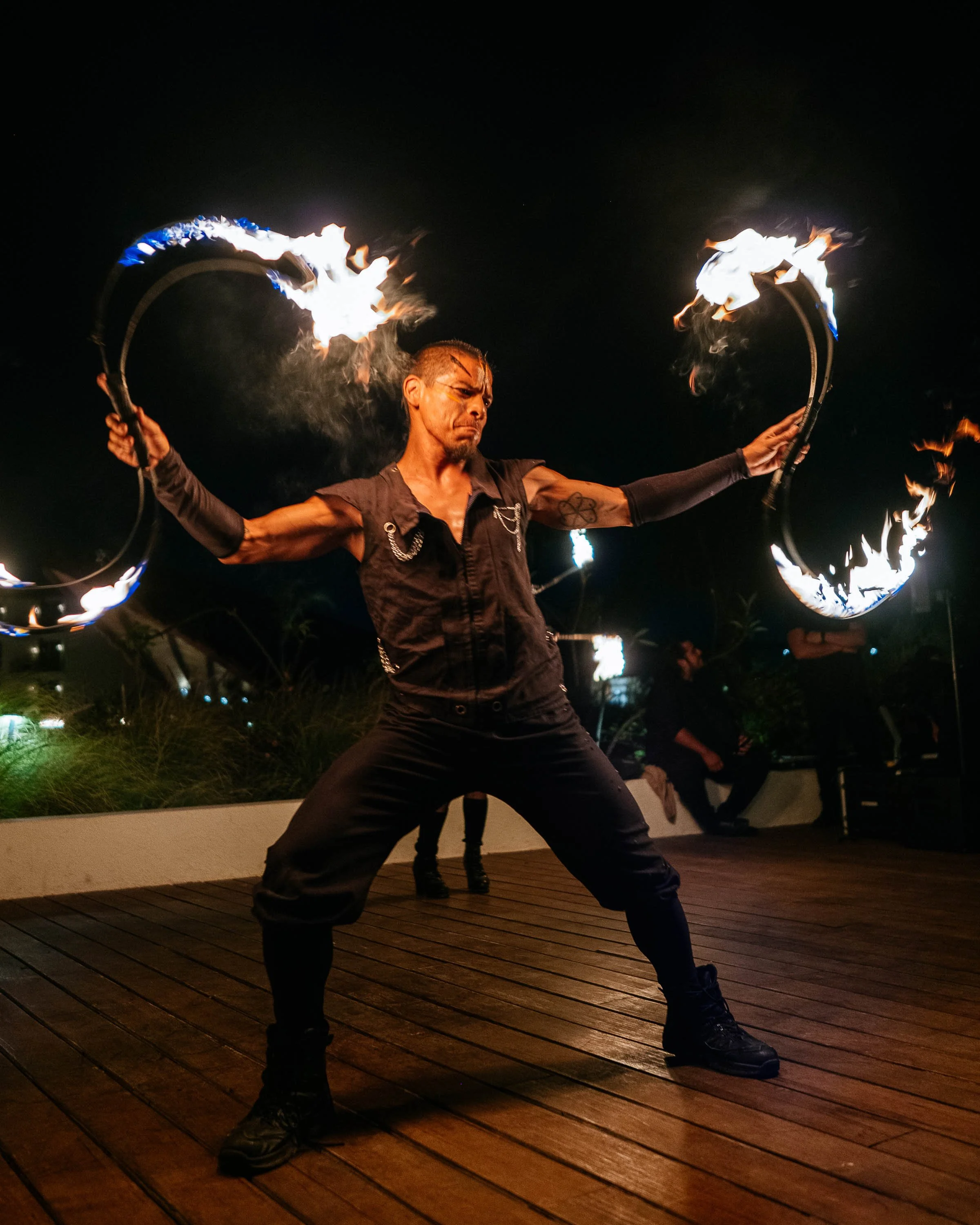 A performer dressed in black, holding two hoops with fire at the ends, creating a fire dance during a nighttime performance.