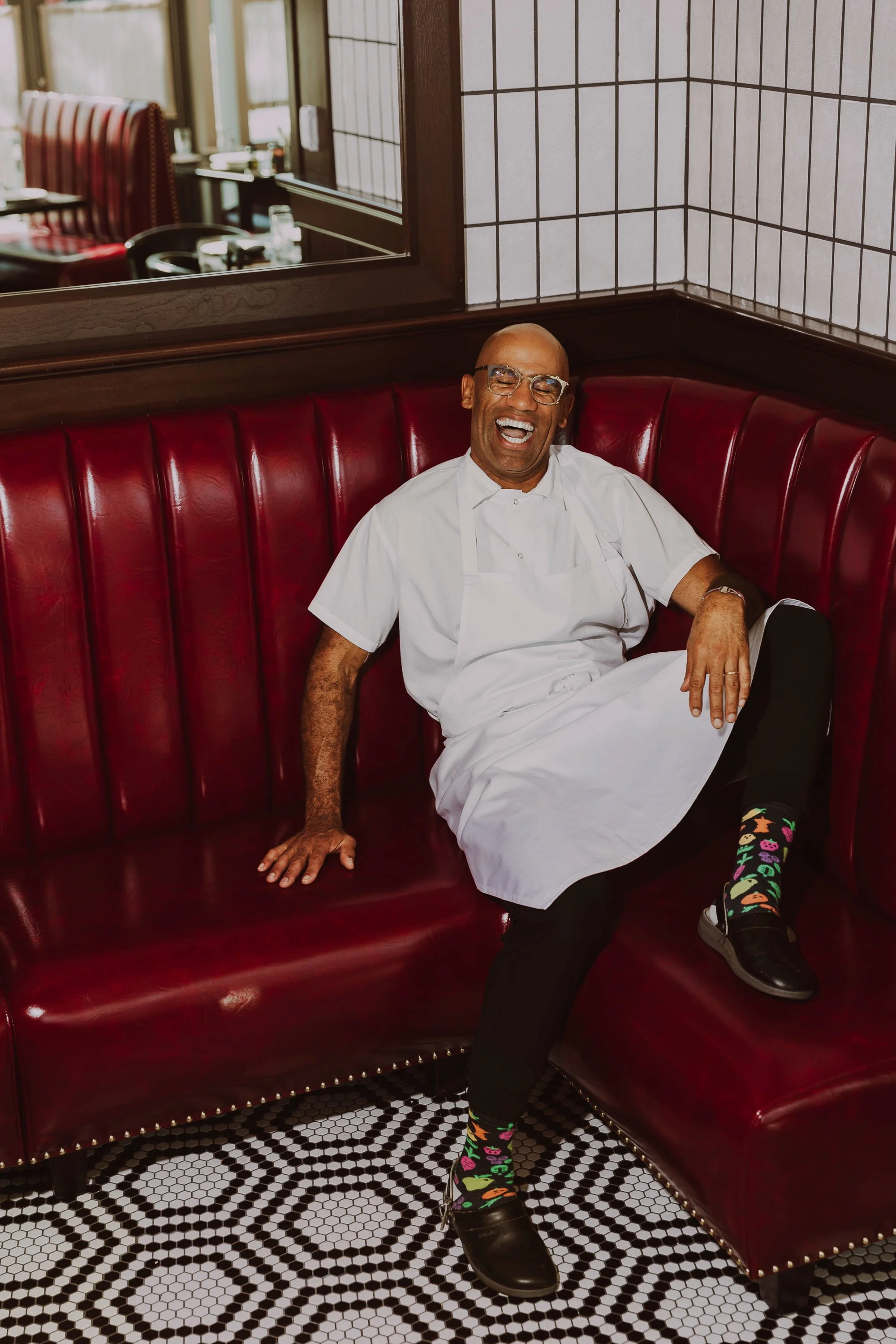 A man in a white chef's coat and glasses sitting on a red leather booth in a restaurant, laughing with colorful socks and black shoes.