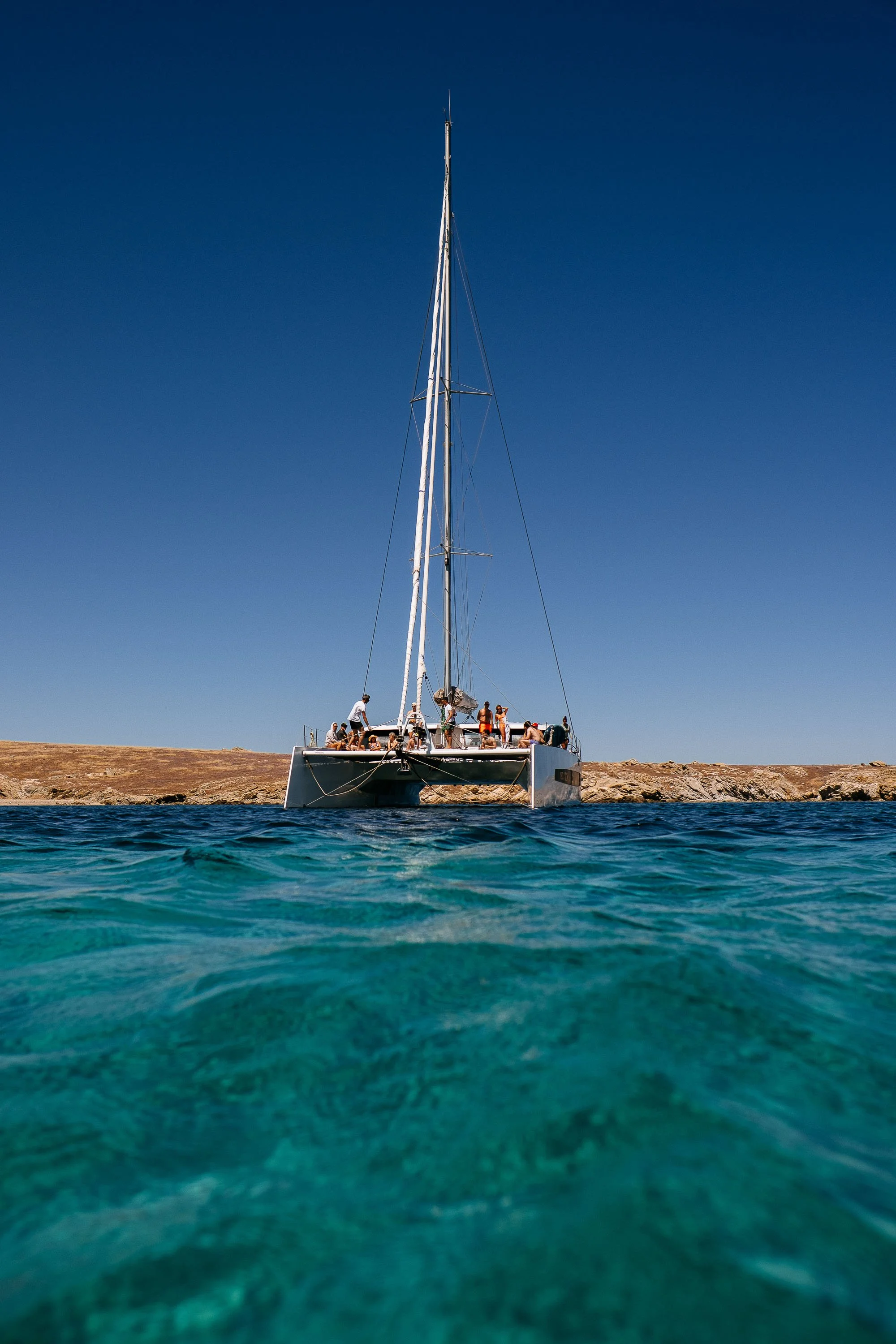A sailboat with people on board floating on clear blue water near a rocky shoreline under a bright blue sky.