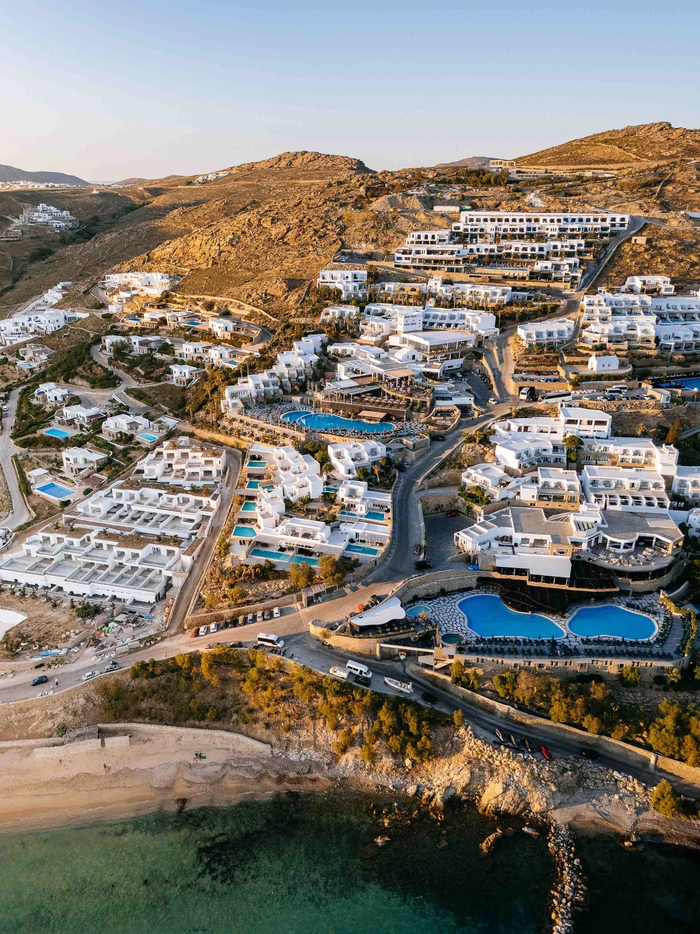 Aerial view of a coastal resort town with white buildings, swimming pools, and winding roads on a hillside during sunset.