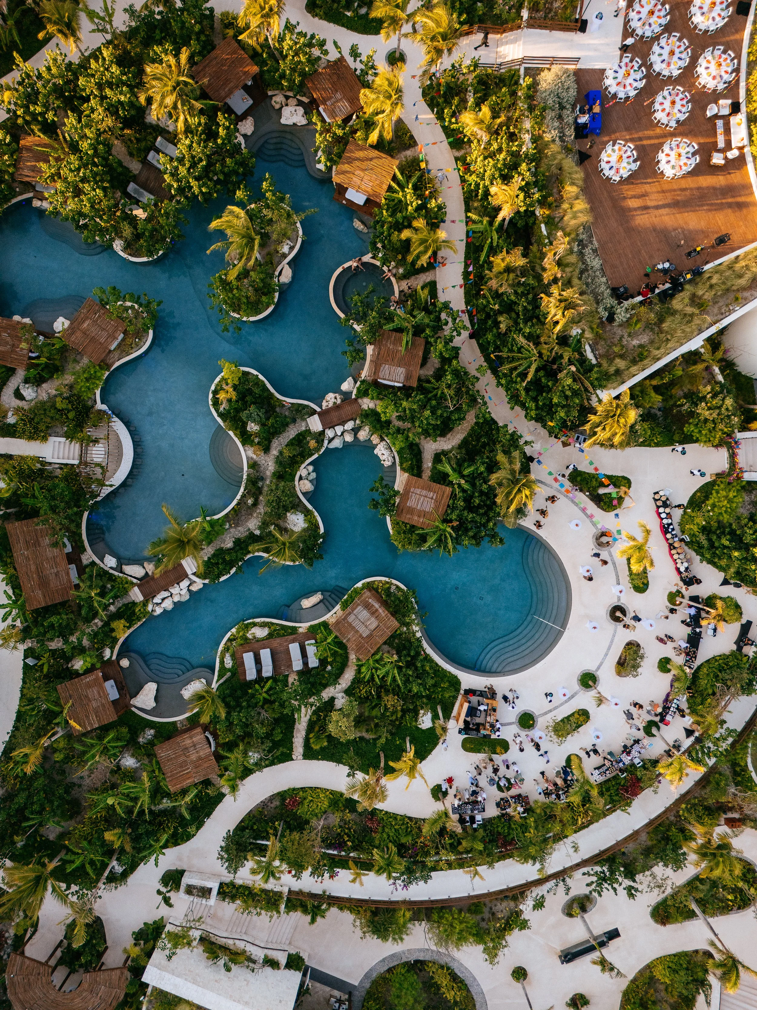 An aerial view of a resort with a large swimming pool surrounded by greenery, small cabanas, lounge chairs, and a deck area with tables and chairs for outdoor dining or events.