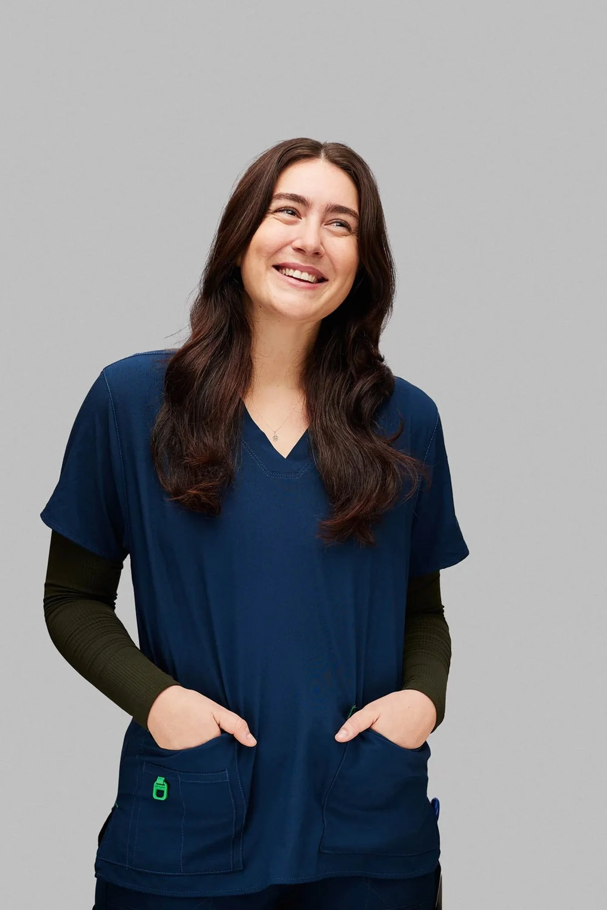 A woman with long dark hair, smiling, wearing medical scrubs with a blue top and black long sleeves underneath, standing against a plain grey background.