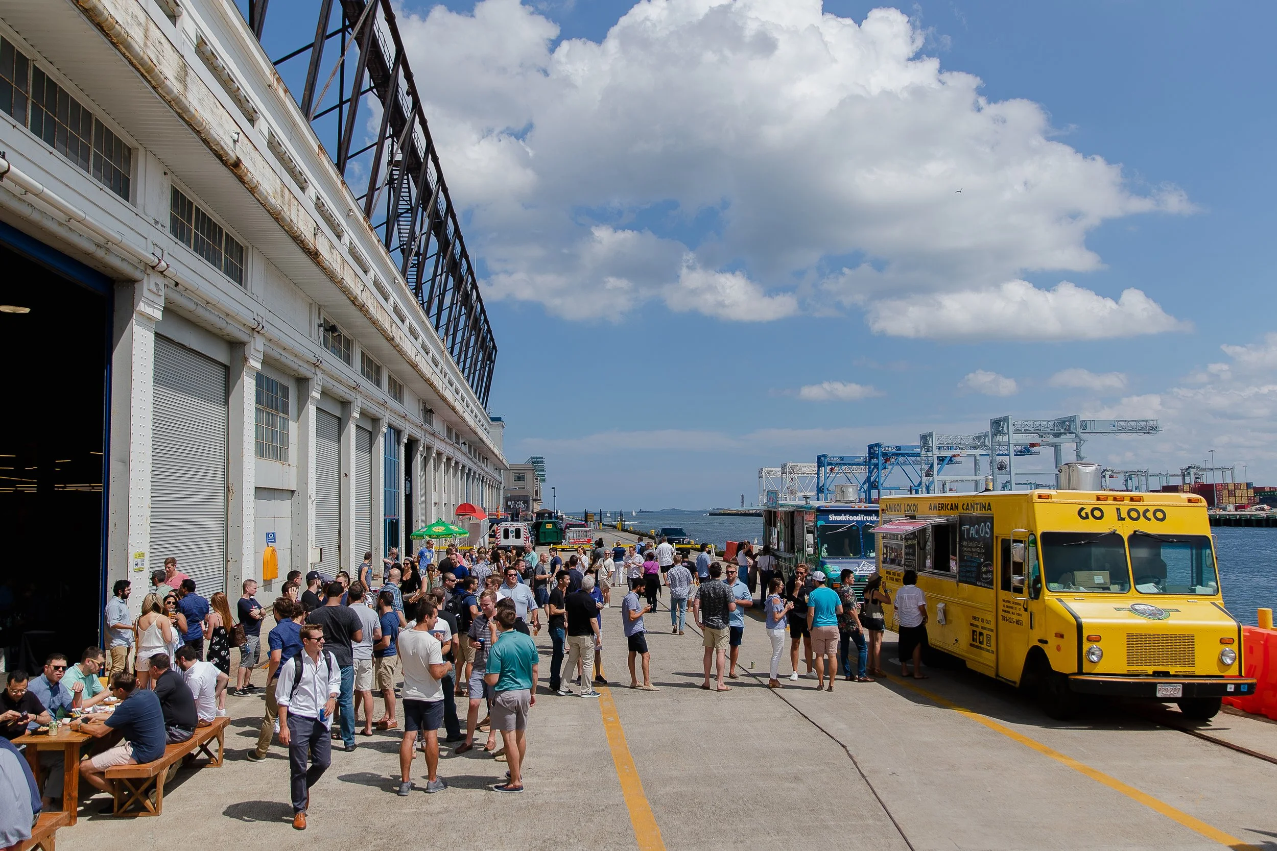 People lined up along a pier near a large building with a view of the harbor, trucks, and cranes in the background on a sunny day with partly cloudy skies.