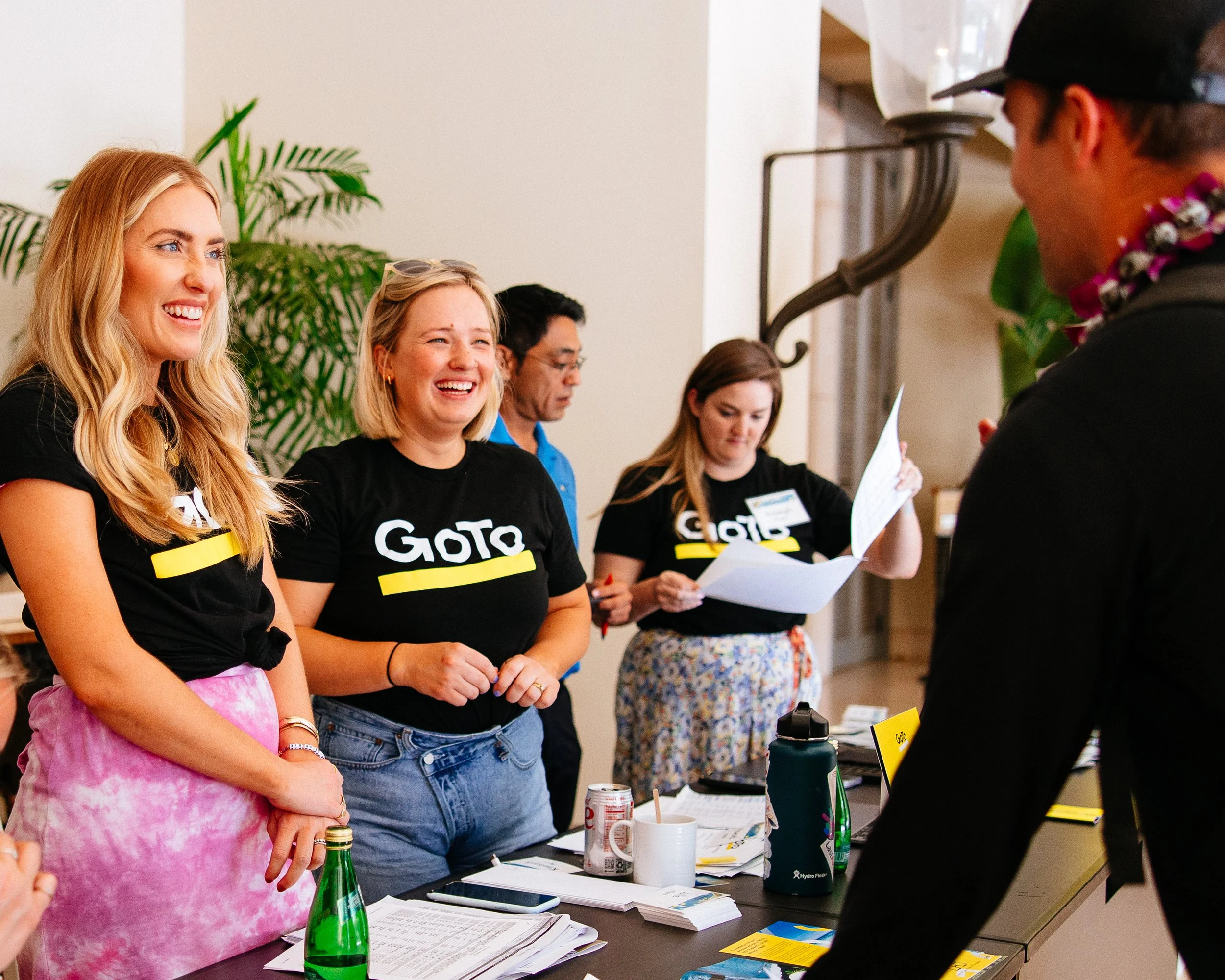 Group of five people at a table, smiling and engaging in conversation, with three women wearing black shirts with 'GOTO' written on them, a man in a blue shirt, and a woman reading a paper; a man in a black cap and sunglasses interacting with them.