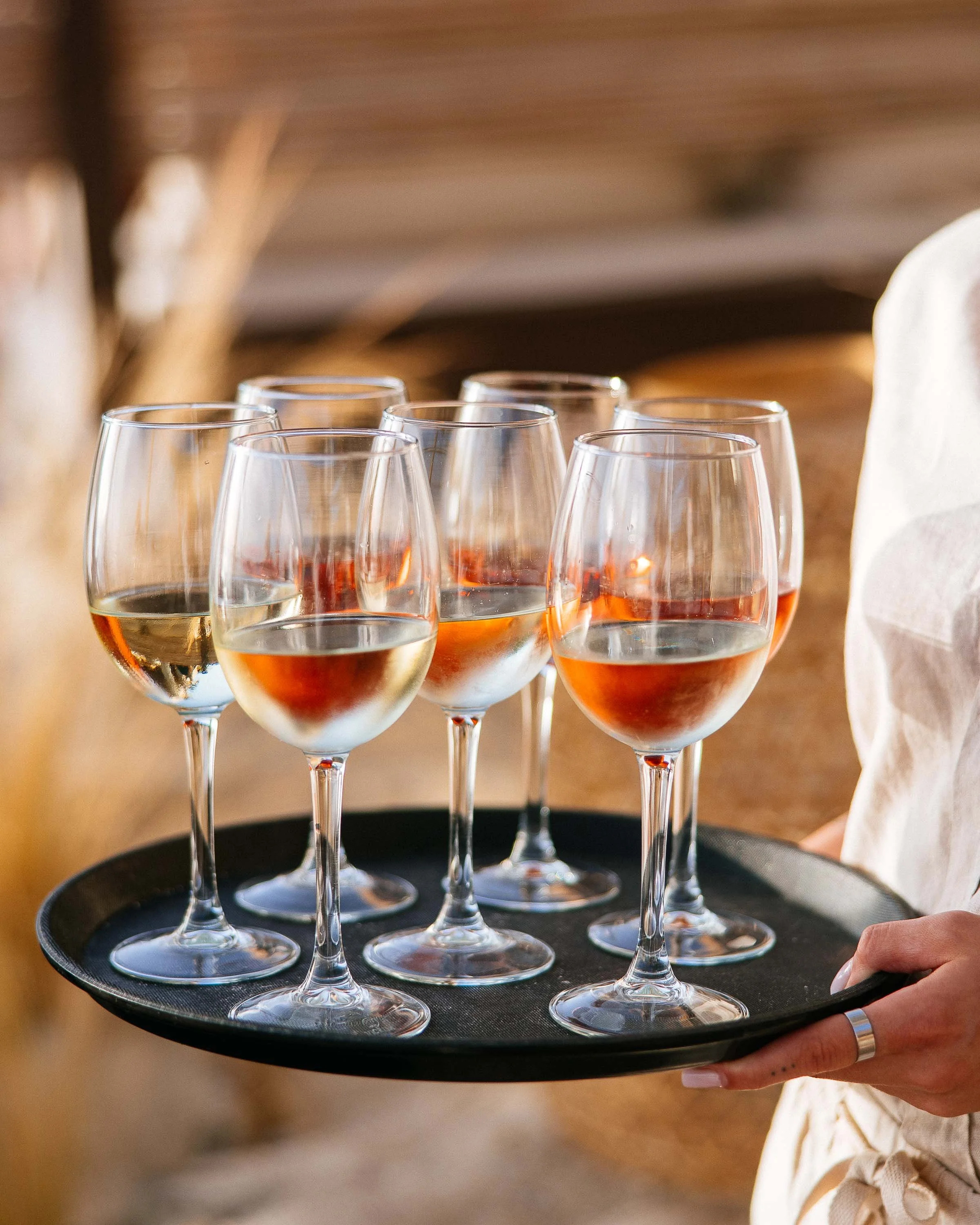A person holding a black tray with six glasses of wine, some filled with white wine and others with rosé, in a setting with warm, natural lighting.