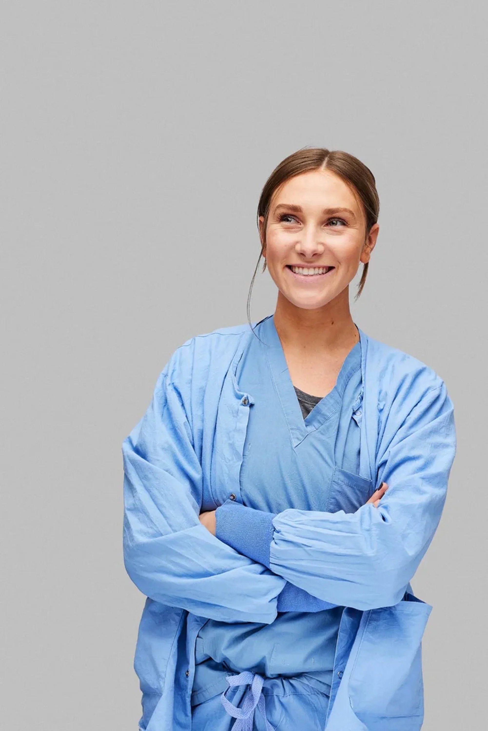 A female healthcare worker in scrubs standing with arms crossed, smiling, against a plain gray background.