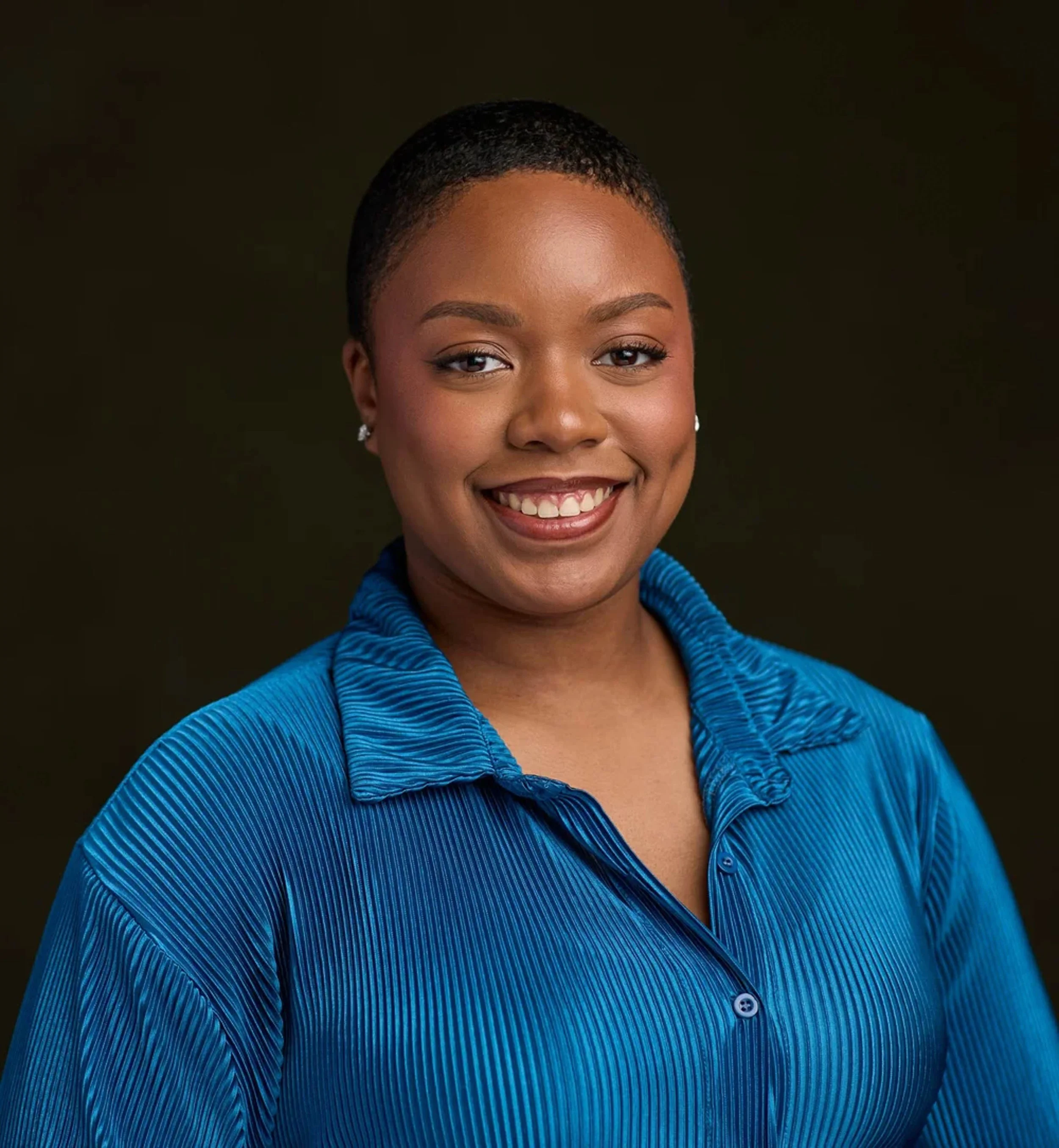 Close-up portrait of a woman with short hair, smiling, wearing a blue textured blazer, against a dark background.