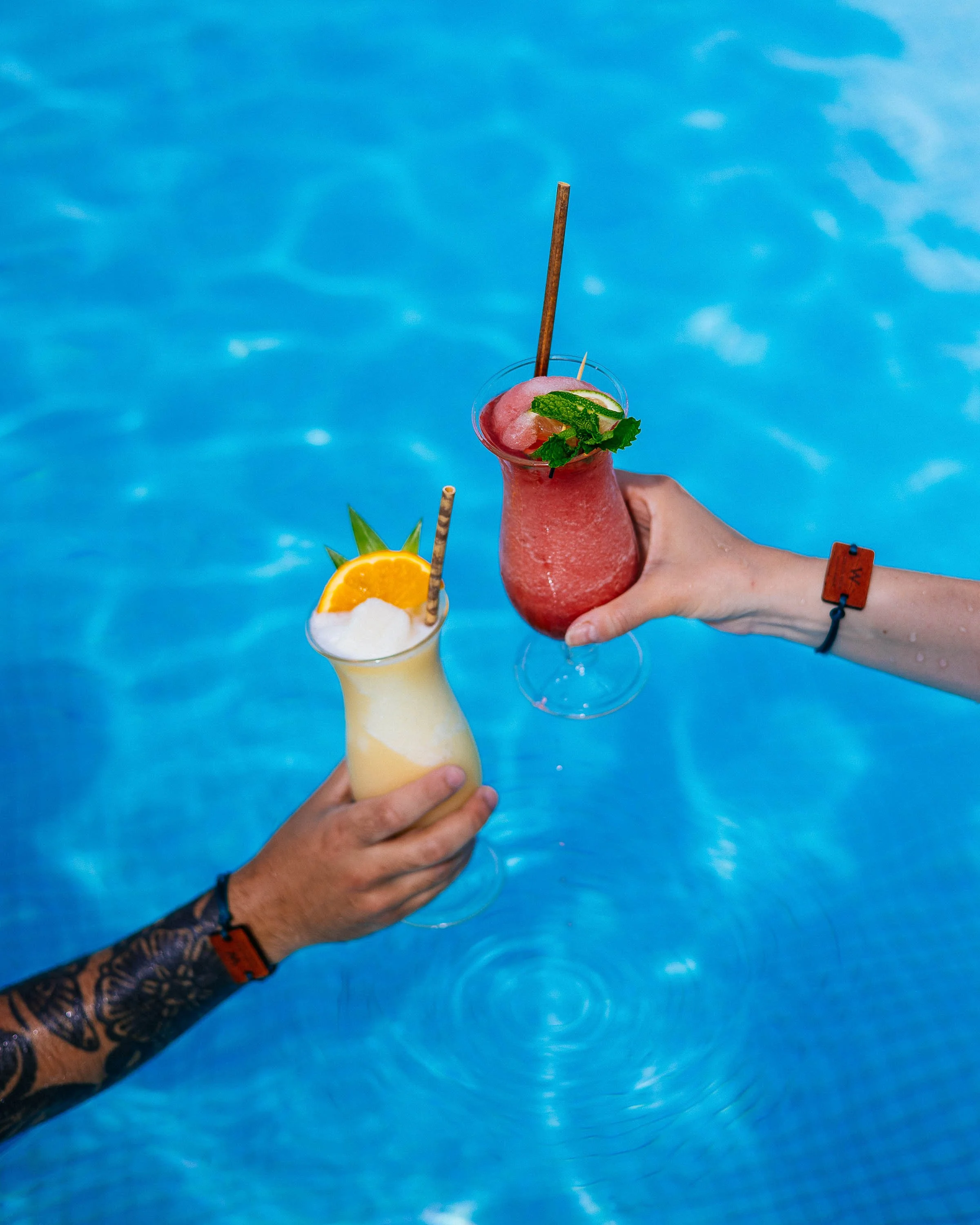 Two hands holding colorful tropical cocktails above a swimming pool. One cocktail is yellow with pineapple and orange garnishes, the other is red with lime and mint garnishes.