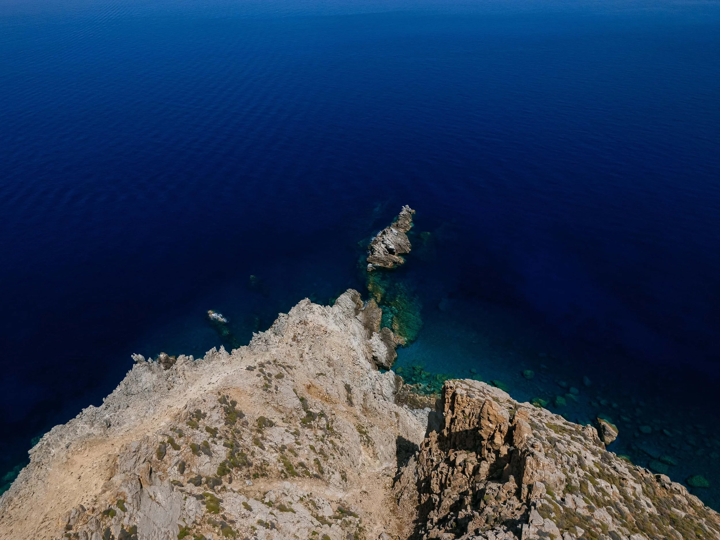 Aerial view of a rocky cliff extending into the deep blue ocean, with clear water around the rocks.