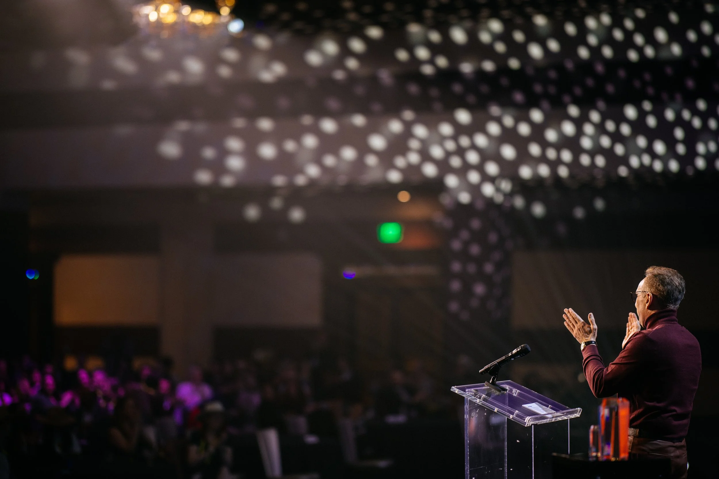A man in a maroon shirt and glasses speaking at a transparent podium during an event, with his hands raised, in front of a dark audience with purple lighting and a decorated ceiling with white circular lights.