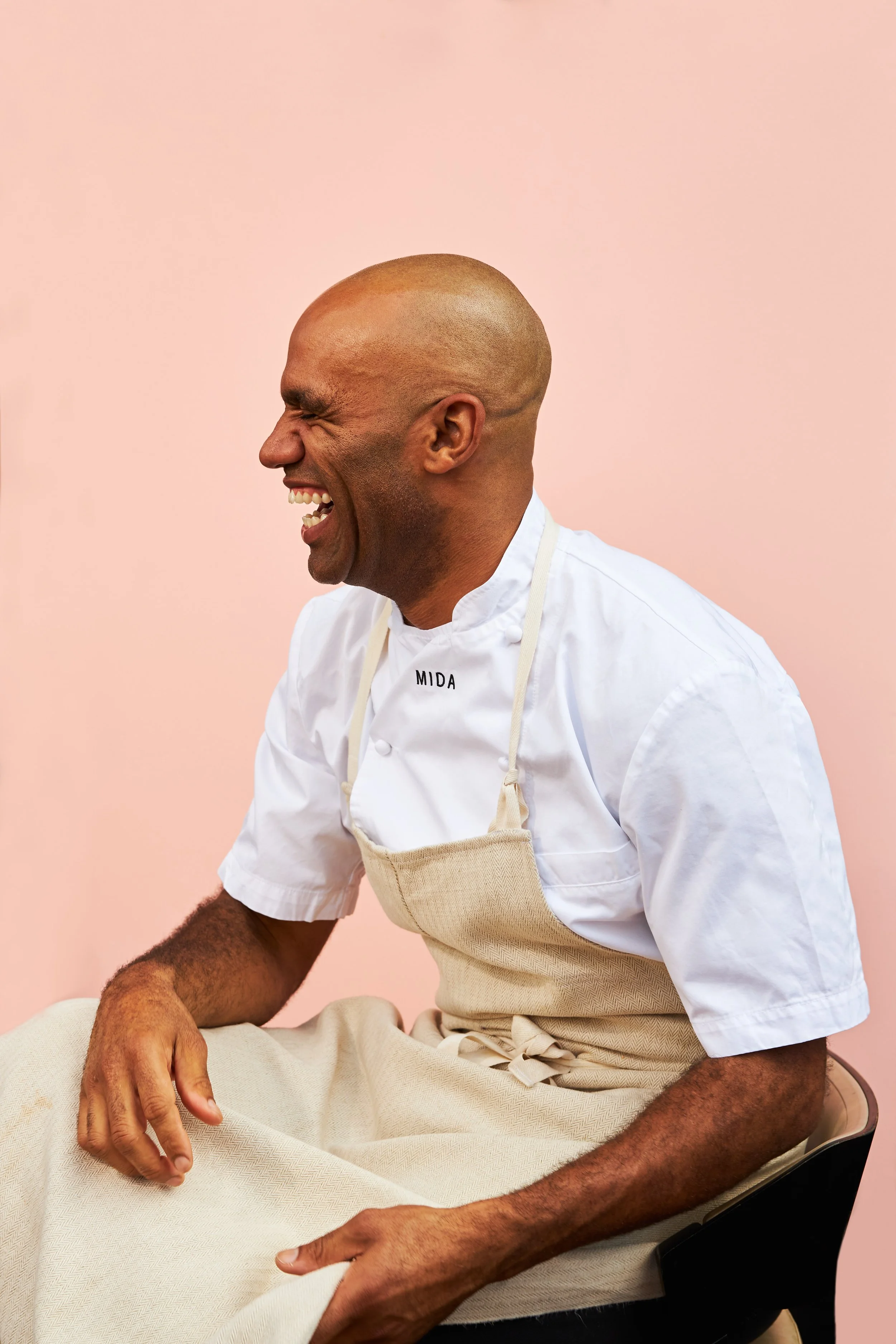 A man in a white chef uniform and beige apron laughing while seated on a black chair against a light pink background.
