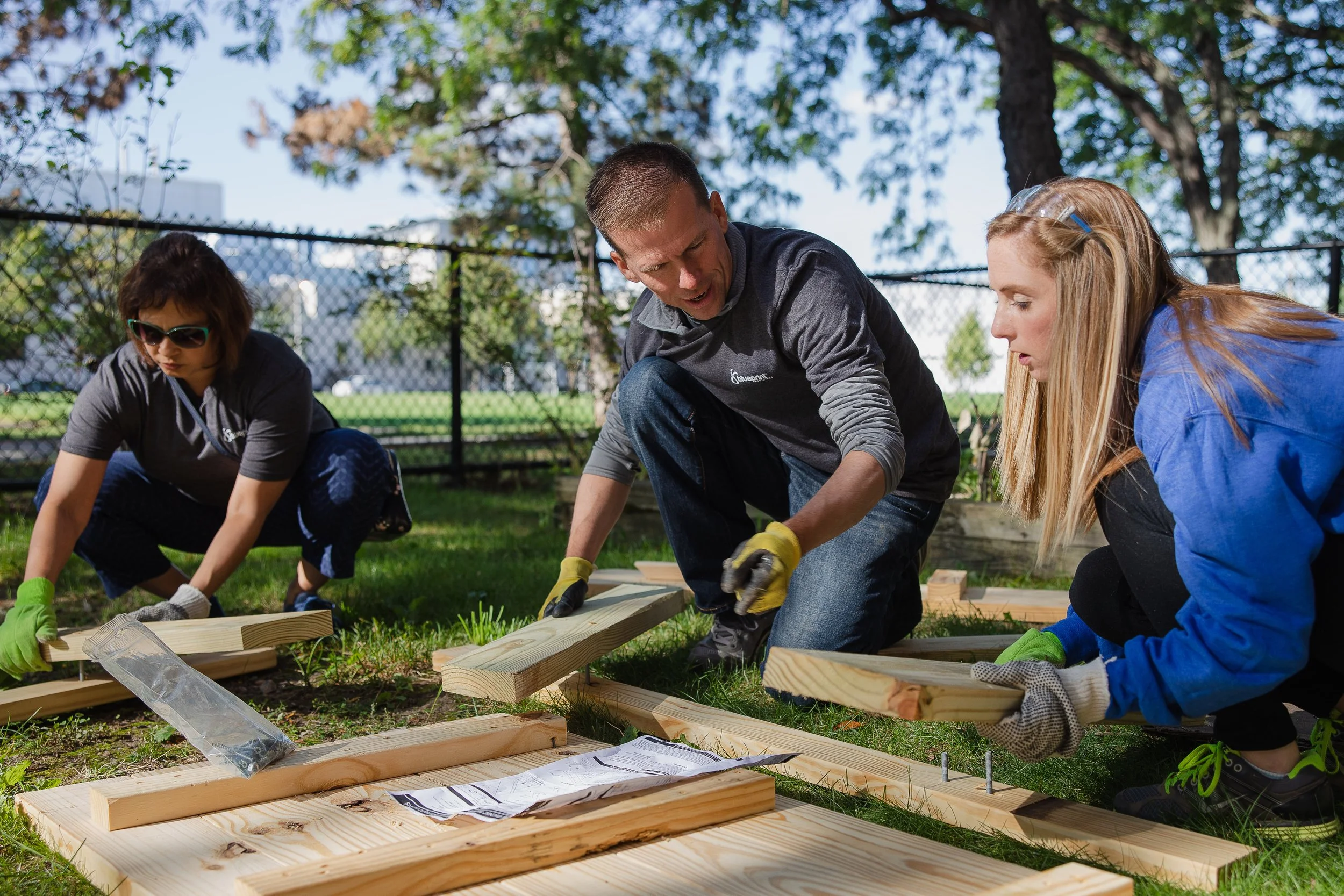 Three people assembling wooden furniture outdoors in a park, wearing gloves and working on wooden planks with a sheet of instructions.