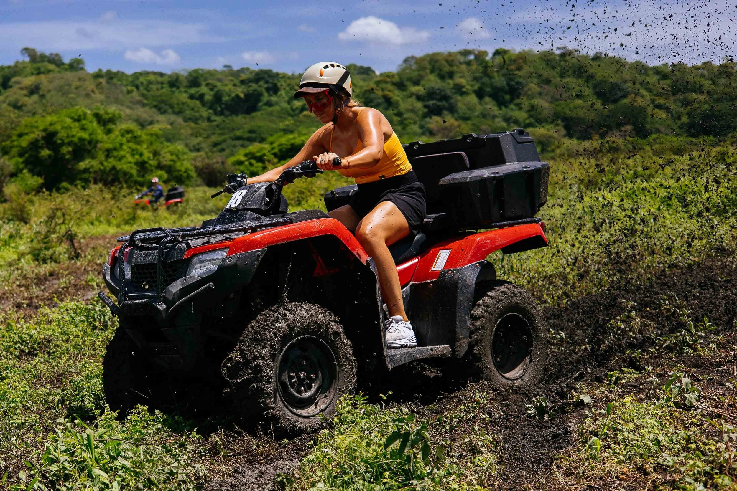 Woman riding a red all-terrain vehicle (ATV) through muddy terrain in a lush green outdoor setting, wearing a helmet and casual athletic clothing.