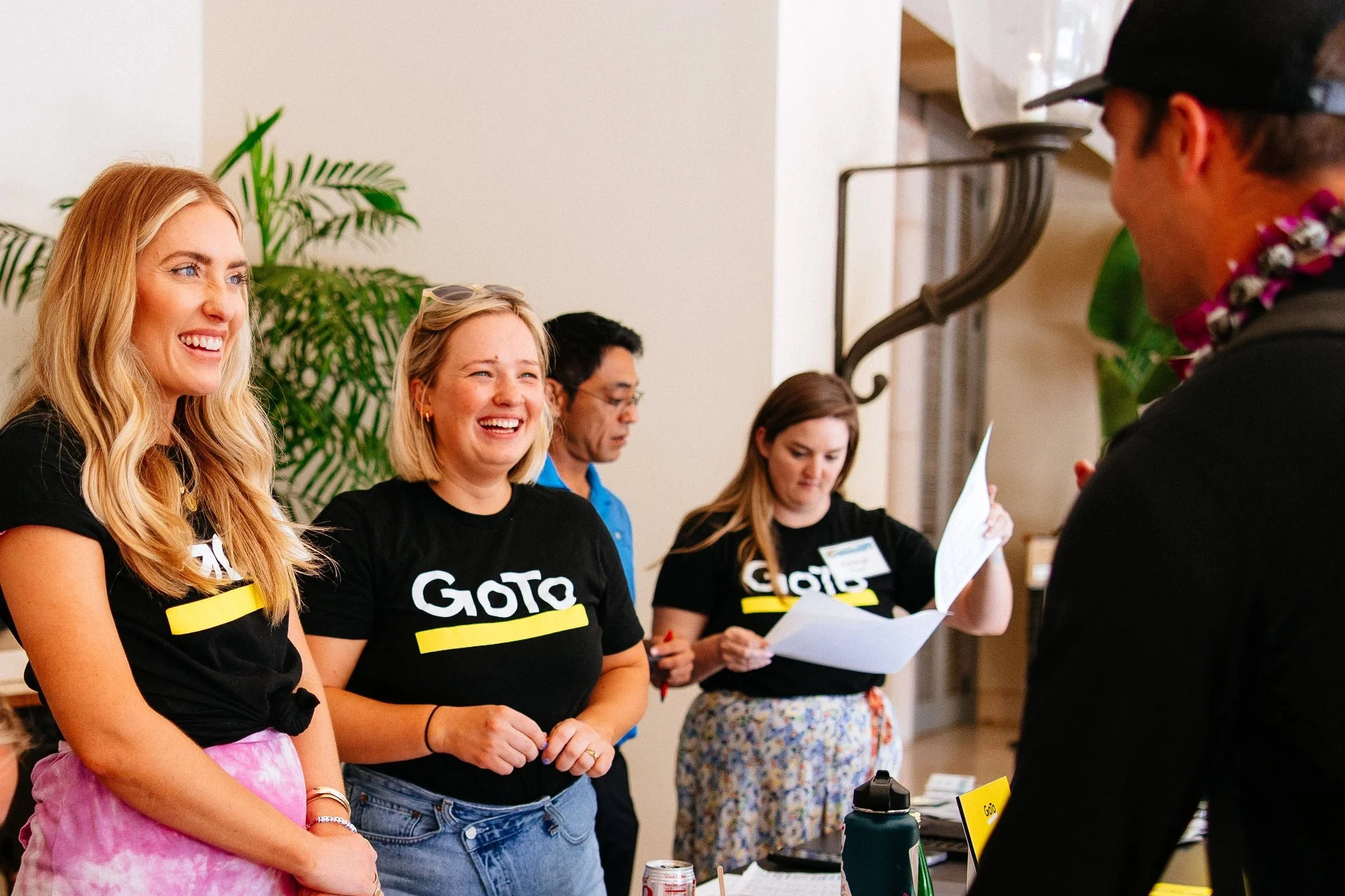 A group of women wearing black t-shirts with 'GOTO' printed on them, standing in a line at an indoor event, smiling and engaging with a person in front of them who is partially visible. One woman has blonde hair and is wearing pink tie-dye shorts, while others have different hairstyles and are holding papers or printed materials.