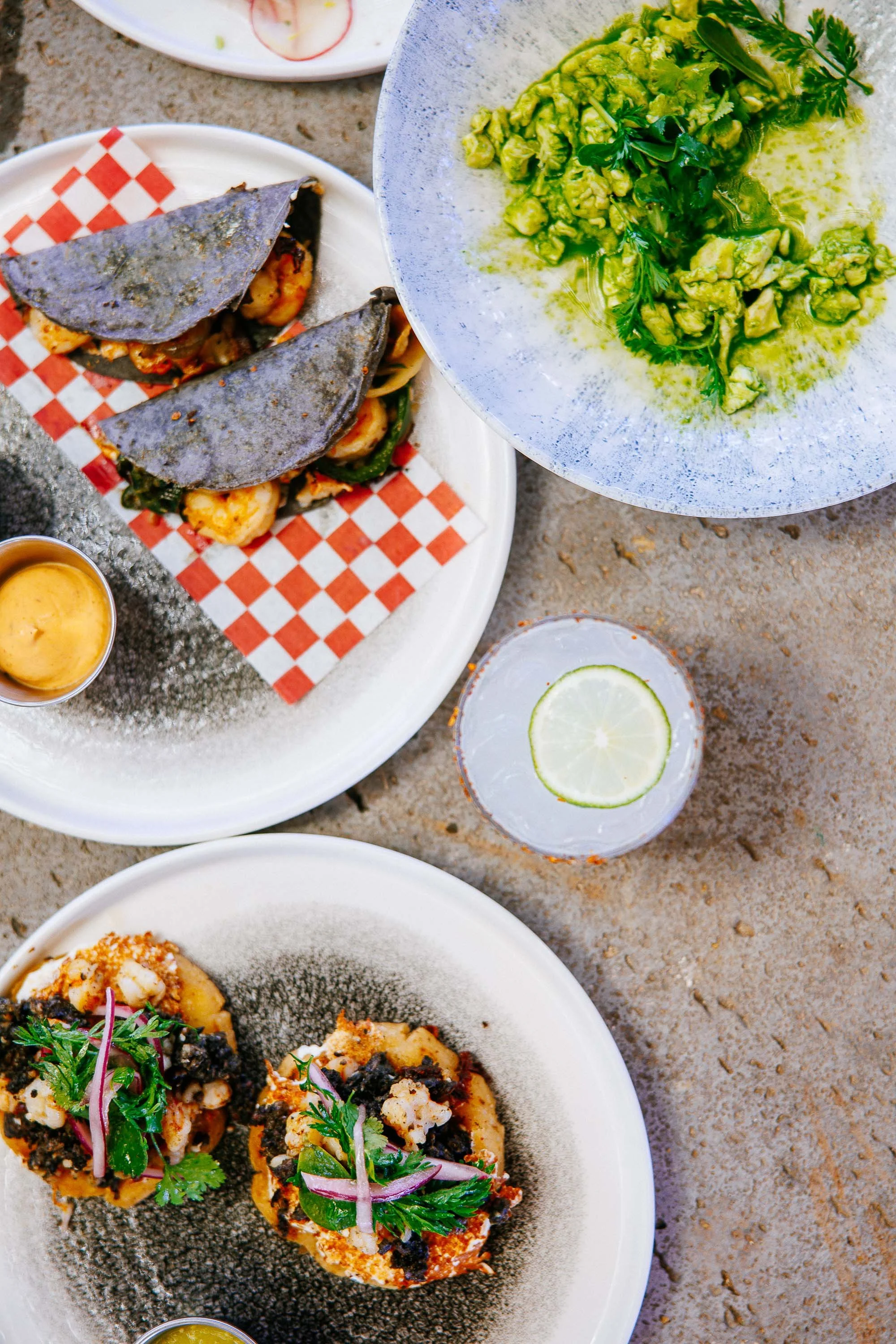 Various Mexican dishes including shrimp tacos with blue corn tortillas, guacamole with cilantro, and two small bowls of sauce on a textured surface.