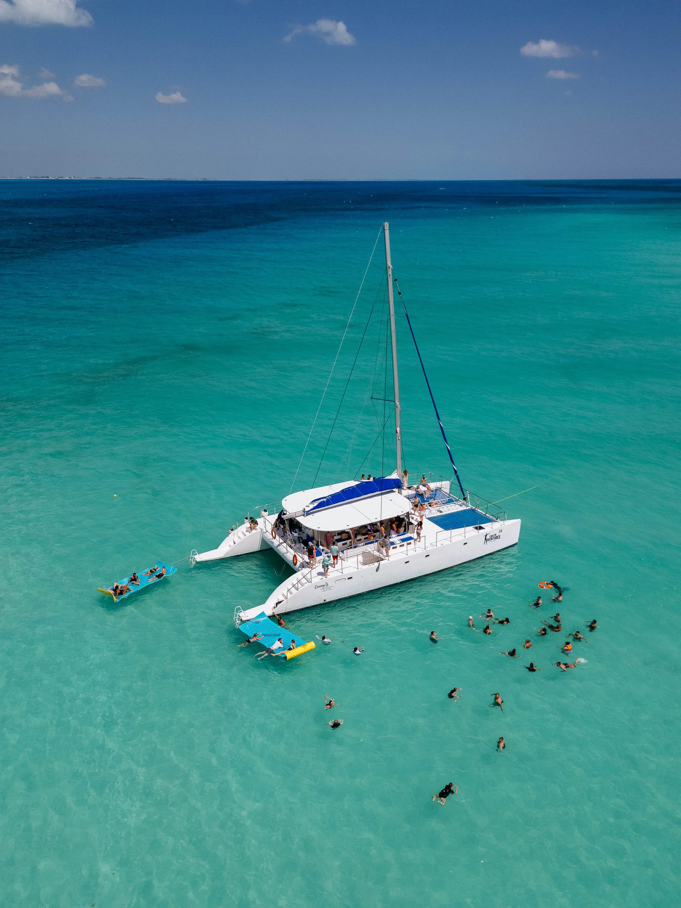 Aerial view of a white yacht with a large group of people on and around it, floating in clear turquoise water with several swimmers and paddleboarders nearby.