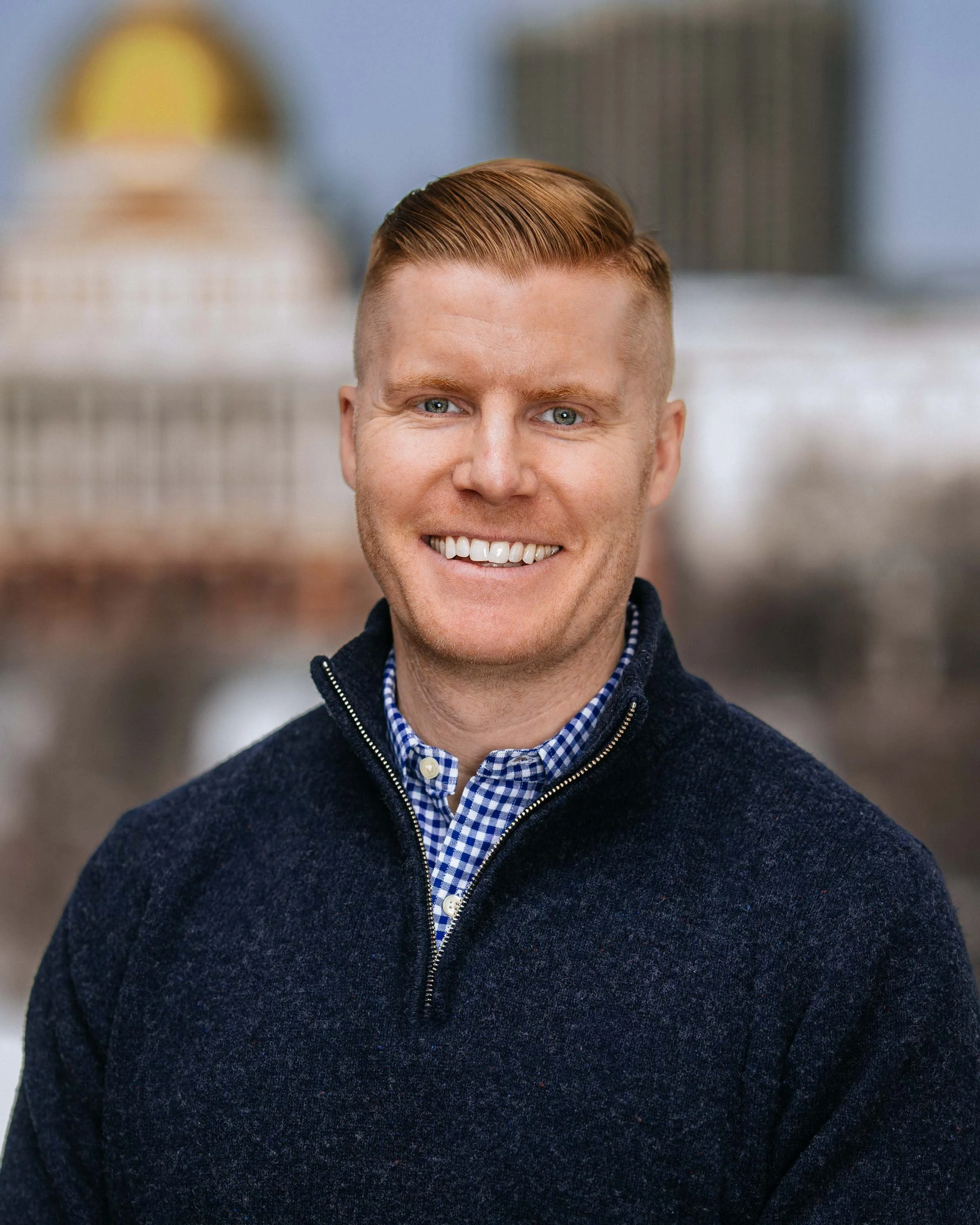 Close-up of a smiling man with short reddish hair, wearing a navy blue zip-up sweater over a blue and white checkered shirt, standing outdoors with blurred background.
