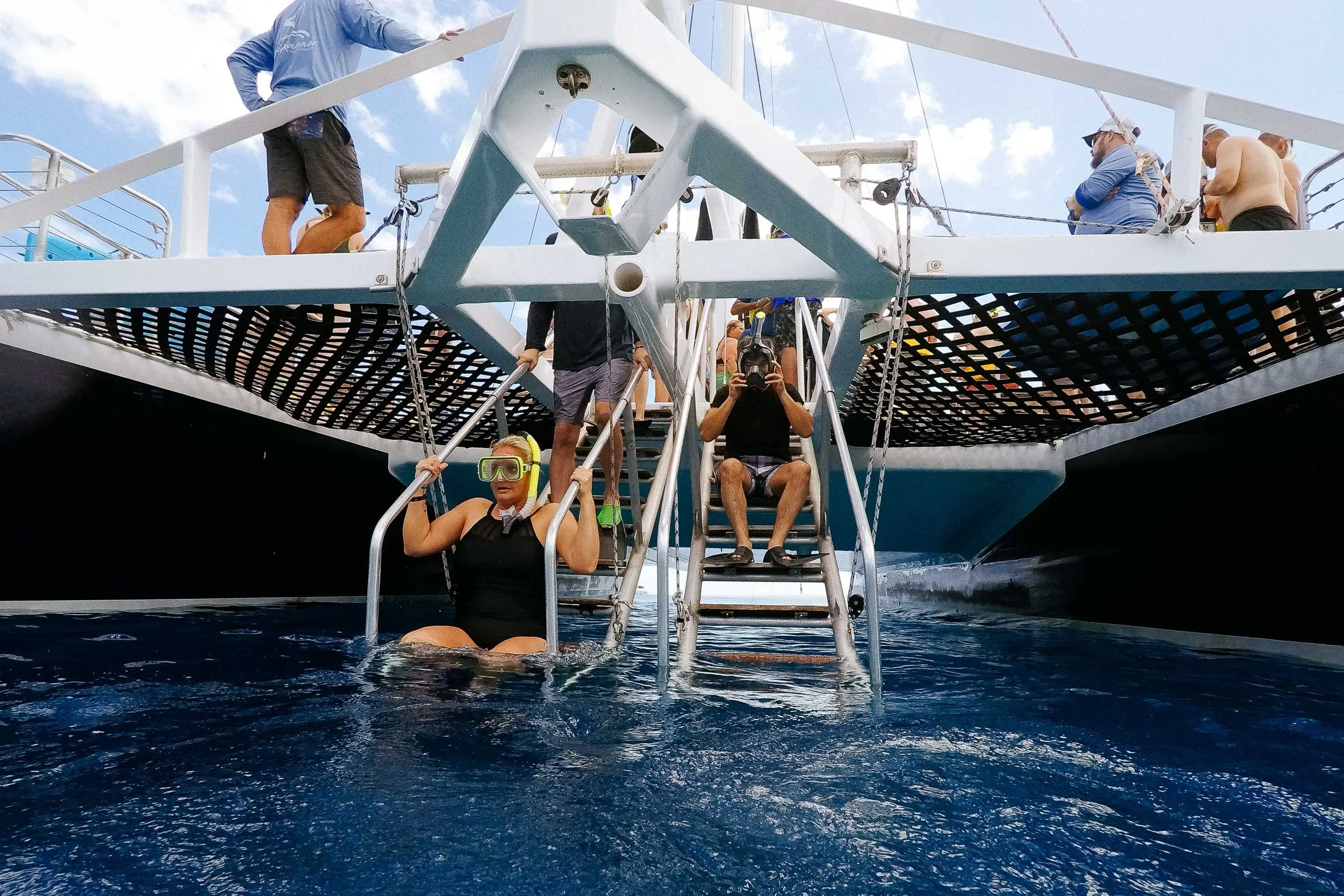 People on a boat, one woman in a black swimsuit with snorkeling gear in the water, two men sitting on the steps of a boat, one with a camera, and others standing or sitting on the boat's deck.