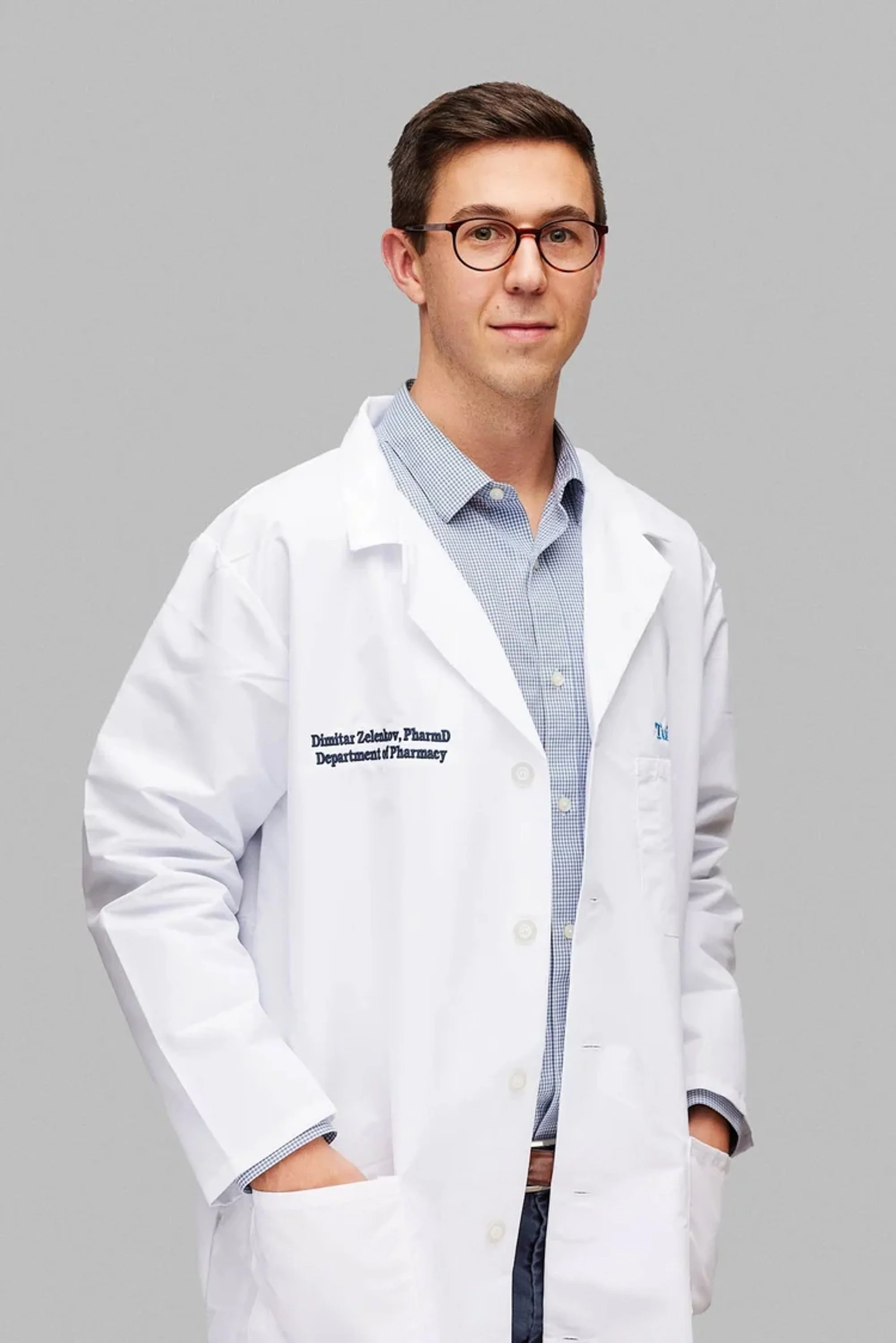 A young male doctor wearing glasses and a white coat with a name tag, standing against a gray background.