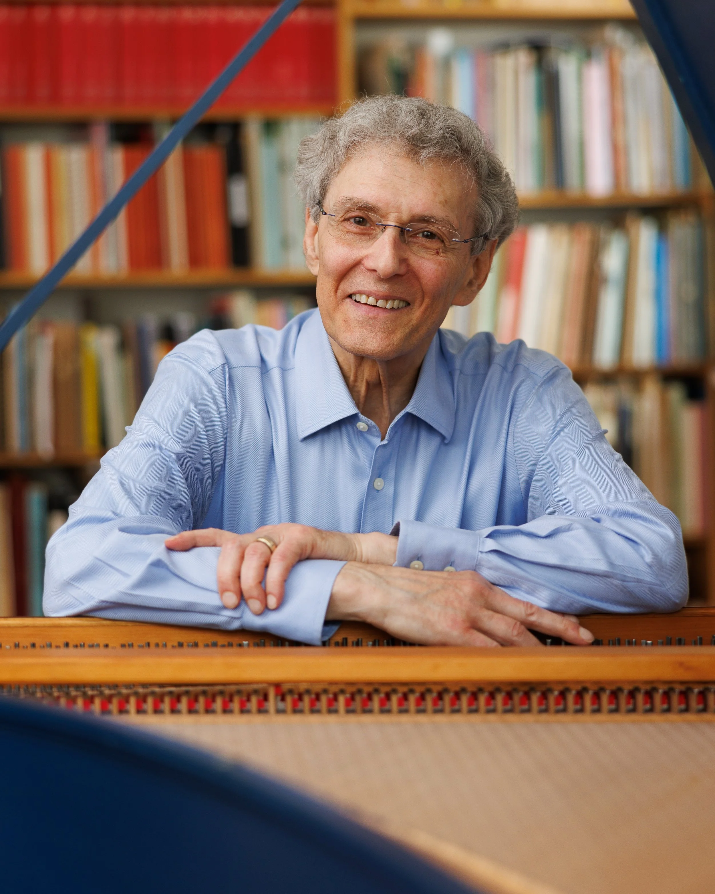 An elderly man with gray hair and glasses smiling, sitting at a piano with a background of bookshelves filled with books.