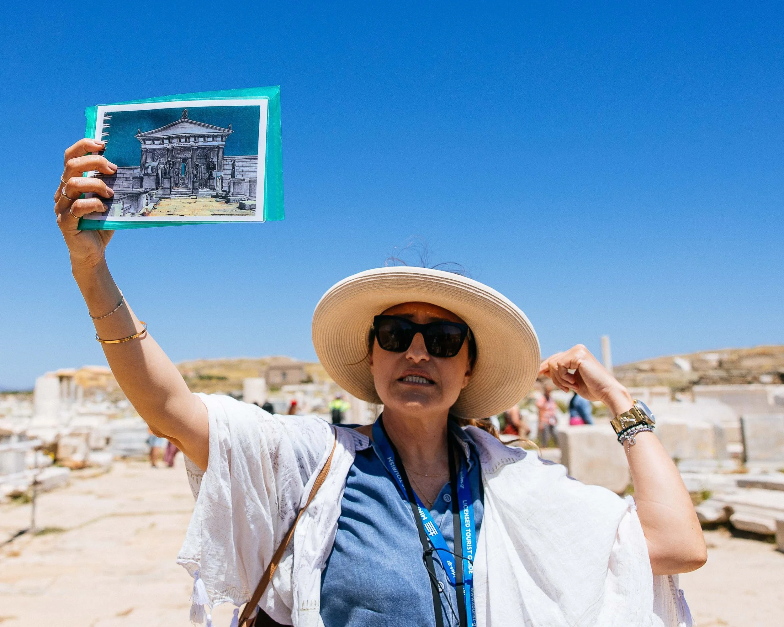 Woman in hat and sunglasses holding a bookmark with an illustrated building image at an archaeological site.