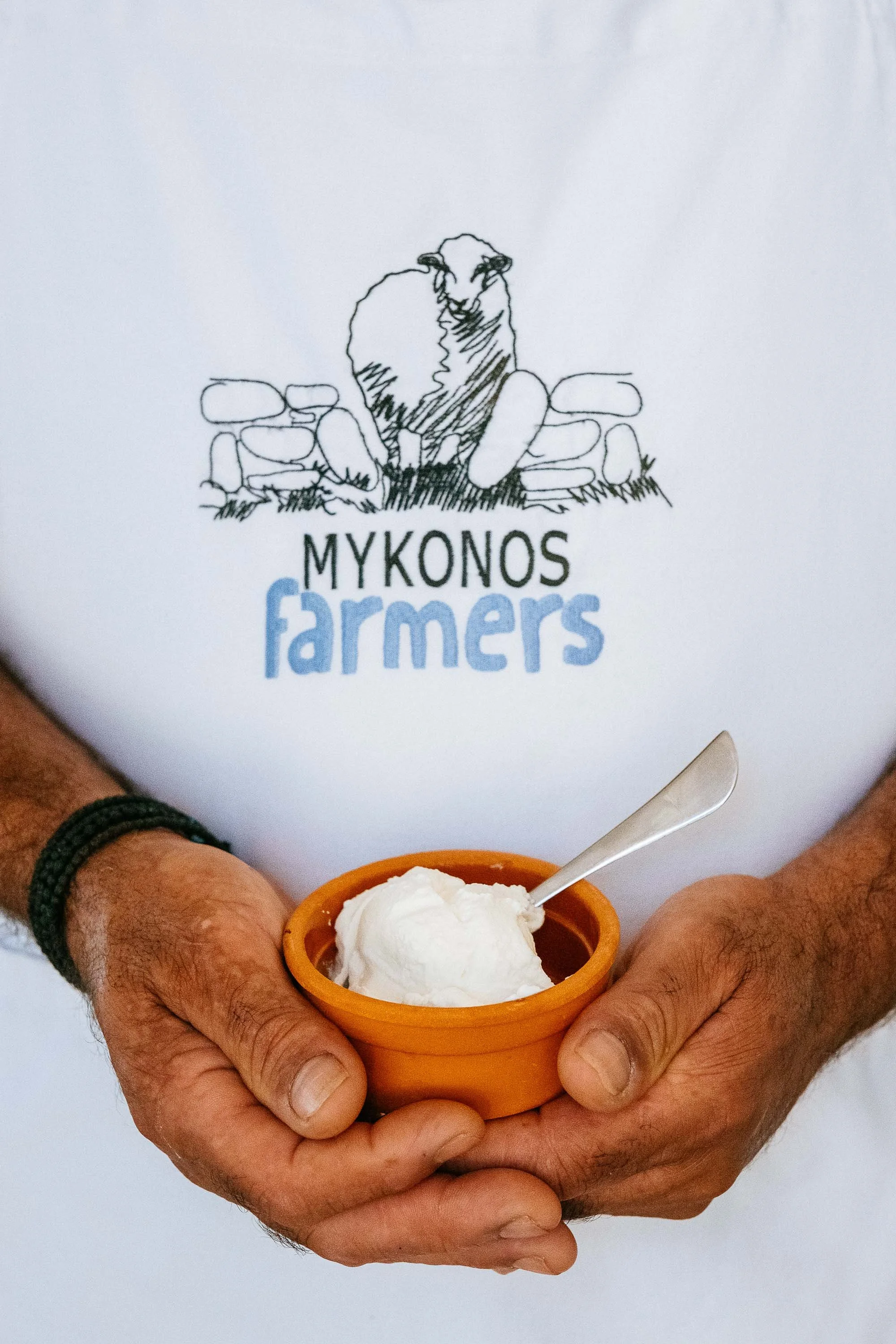 Person wearing a white shirt with a Mykonos farmers logo holding an orange cup of yogurt with a spoon.