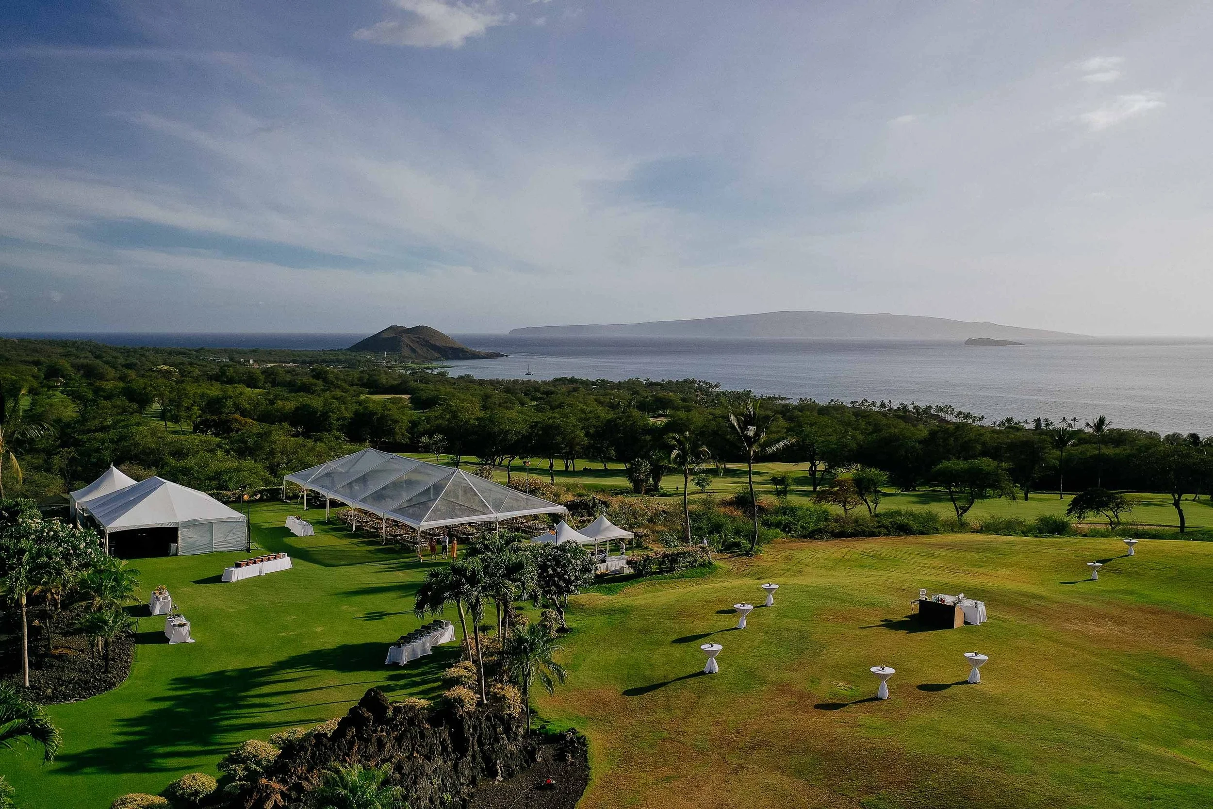 Scenic view of a wedding venue set up on a grassy area near the ocean, with white tents, tables, and scattered seating, overlooking a distant island and body of water under a partly cloudy sky.