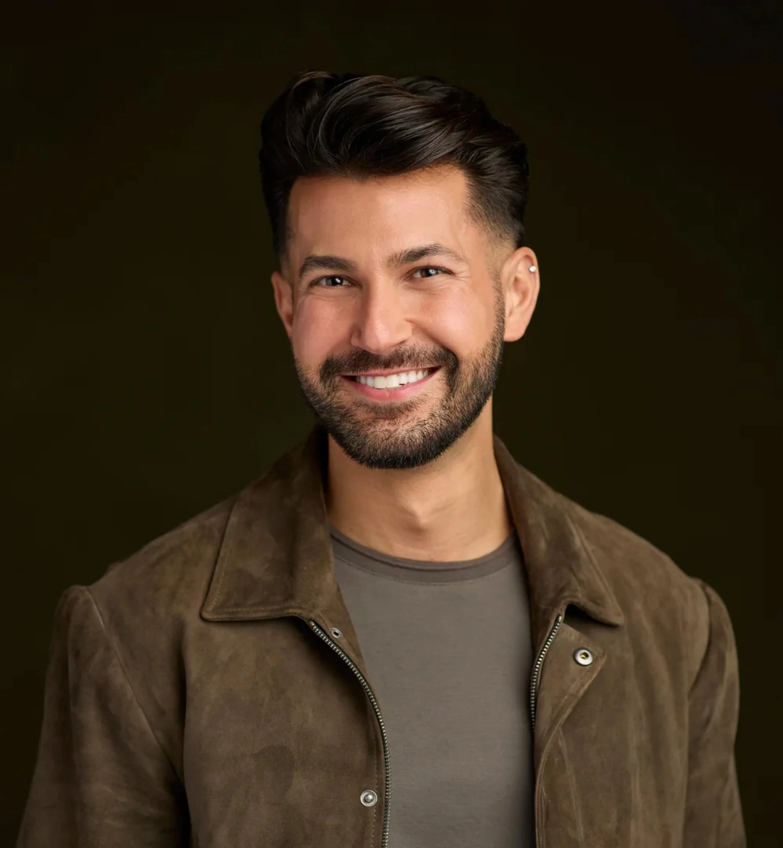 Close-up of a smiling man with dark hair, a beard, and an earring, wearing a brown jacket over a gray shirt, against a dark background.