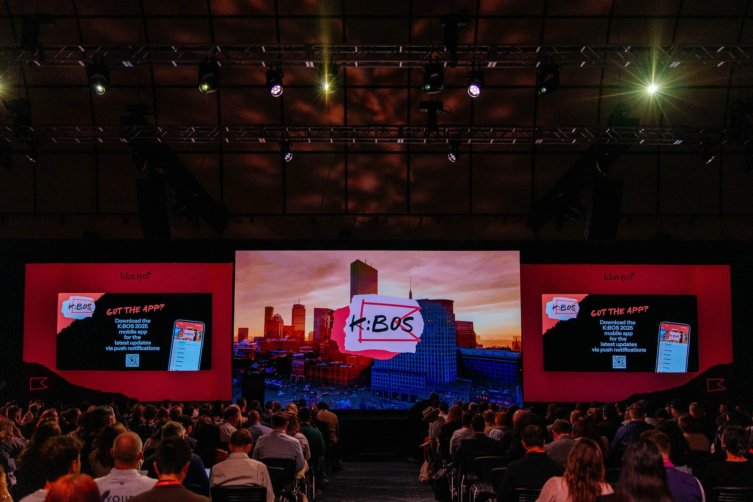 A large conference or presentation event with an audience, and a stage featuring multiple display screens. The main screen shows a city skyline with a sign reading 'K: BOS' and promotional content for the K: BOS 2025 mobile app. The audience is seate