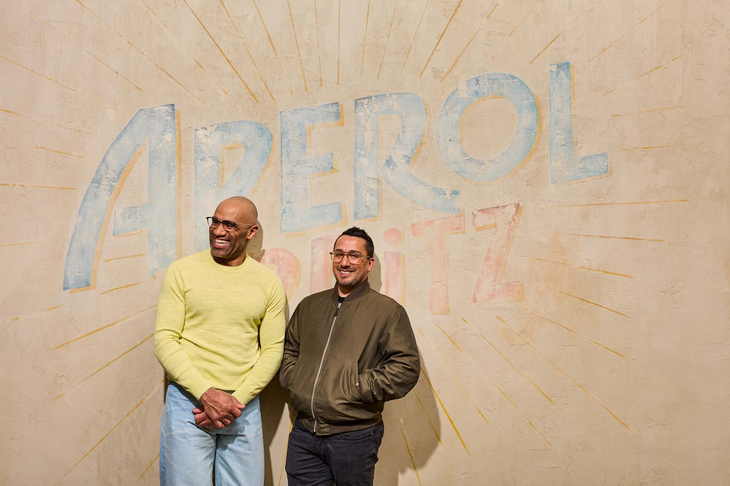 Two men standing in front of a painted wall with the word 'abroad' and rays emanating from it. One man is bald and wearing glasses and a yellow sweater, smiling. The other man has short dark hair, glasses, and a green jacket, smiling.