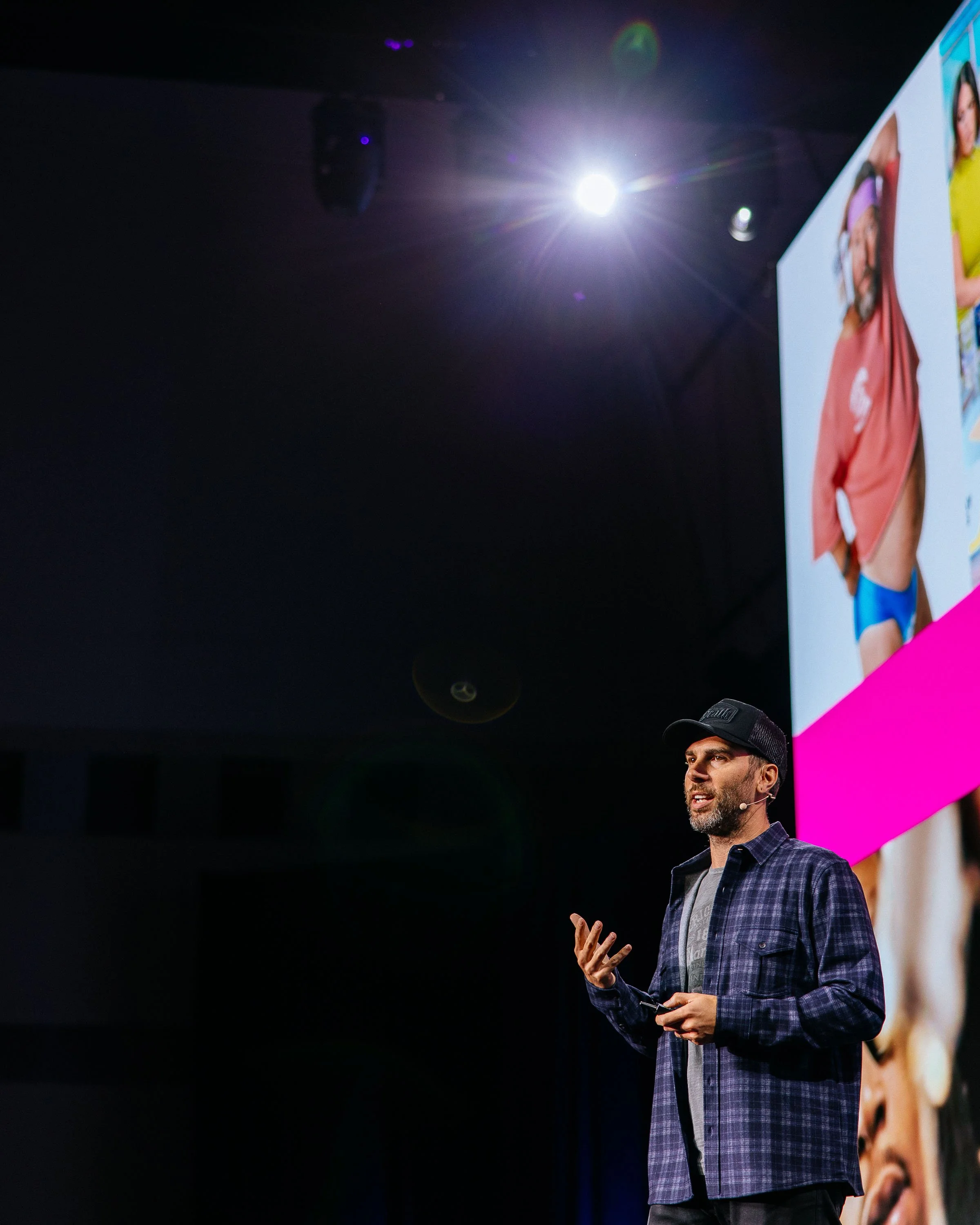 A man wearing a black baseball cap, a plaid shirt, and jeans is giving a presentation on stage. He is speaking and gesturing with his right hand, holding a small object in his left hand. In the background, there is a large screen displaying an image 