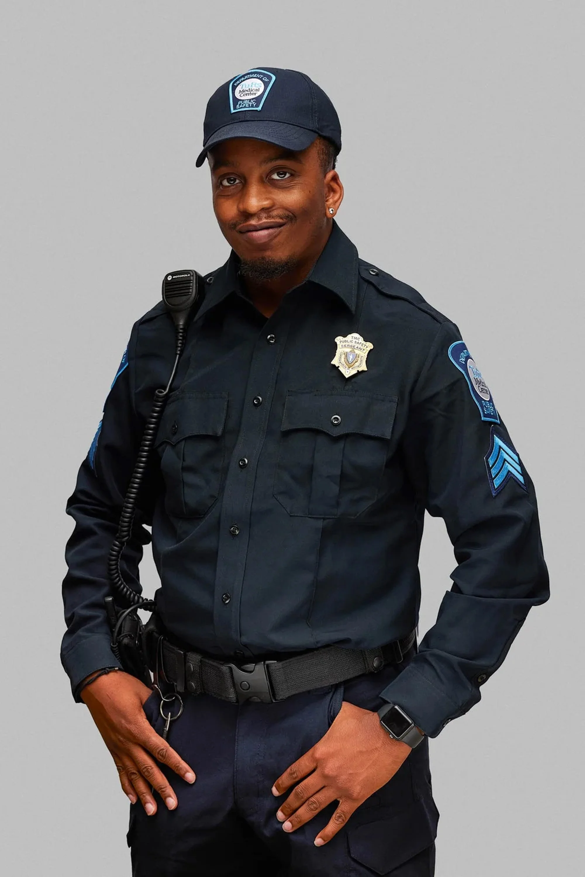 A male police officer in a dark uniform, wearing a cap with a badge, standing against a plain light grey background.