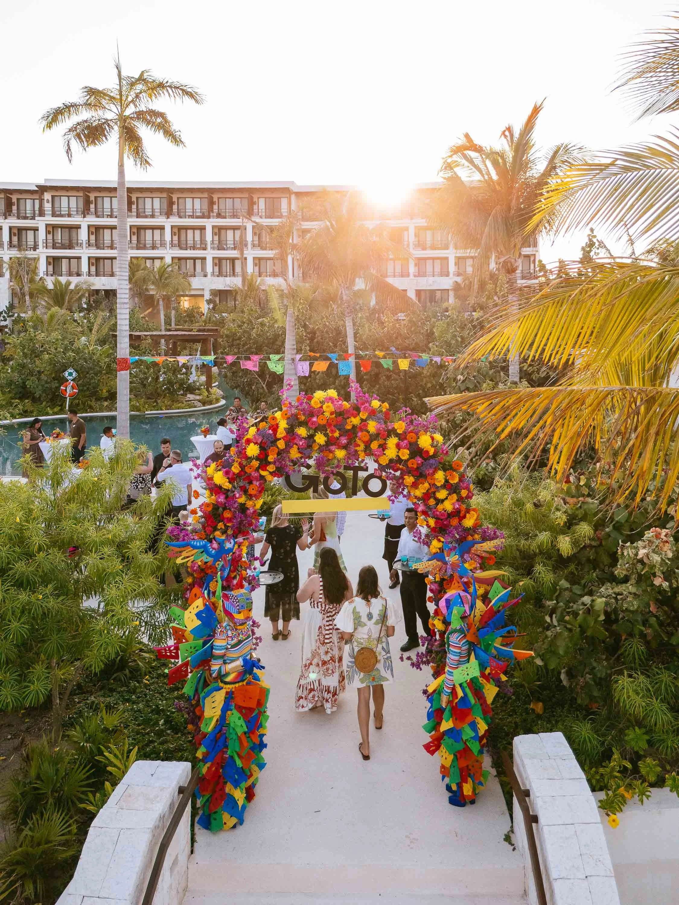 People walking through a tropical outdoor resort area decorated with colorful flowers and banners, near a swimming pool, with a hotel building and palm trees in the background.