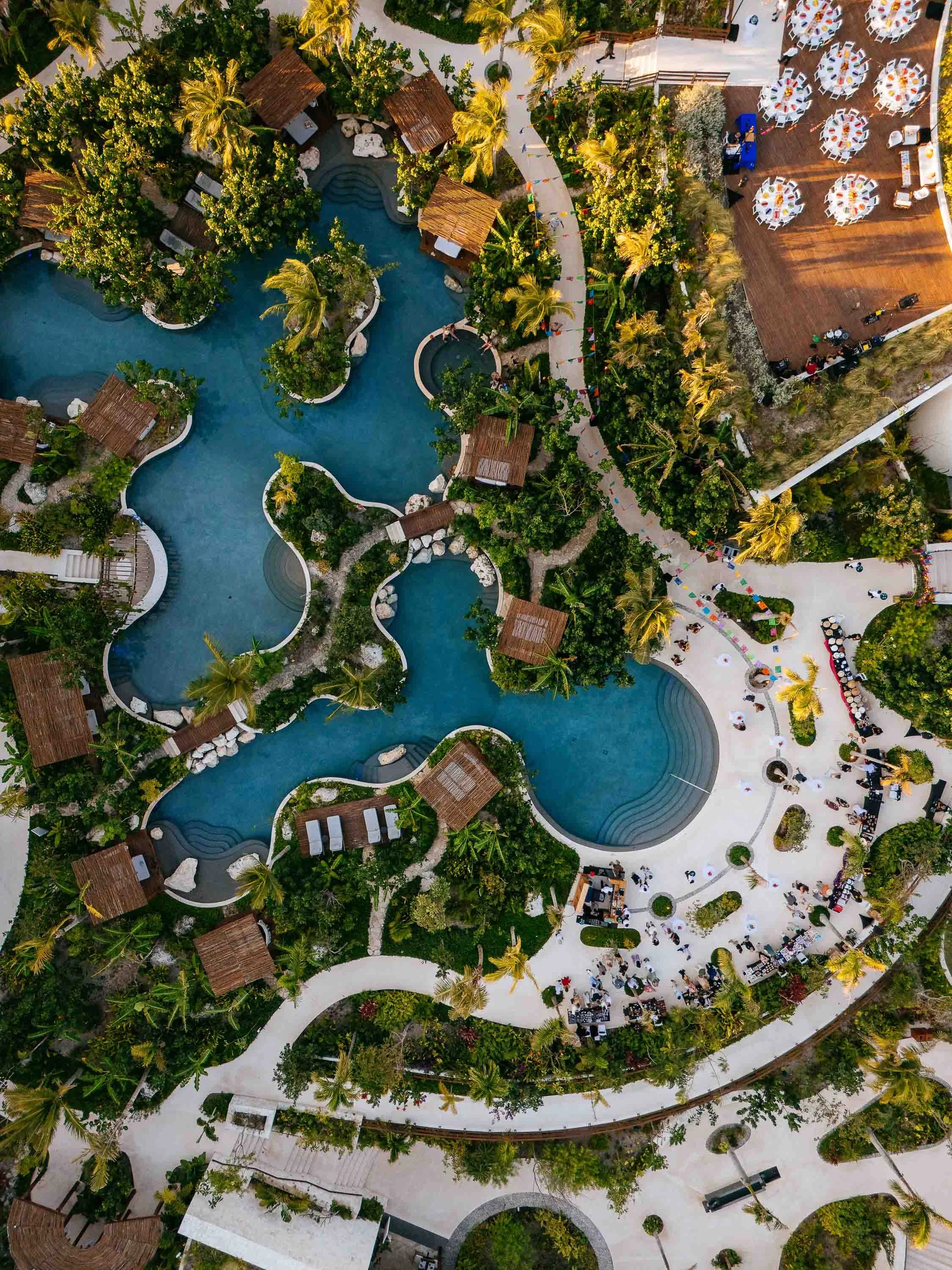 Aerial view of a resort with swimming pools, surrounded by palm trees, walkways, and lounging areas with tables, chairs, and umbrellas.
