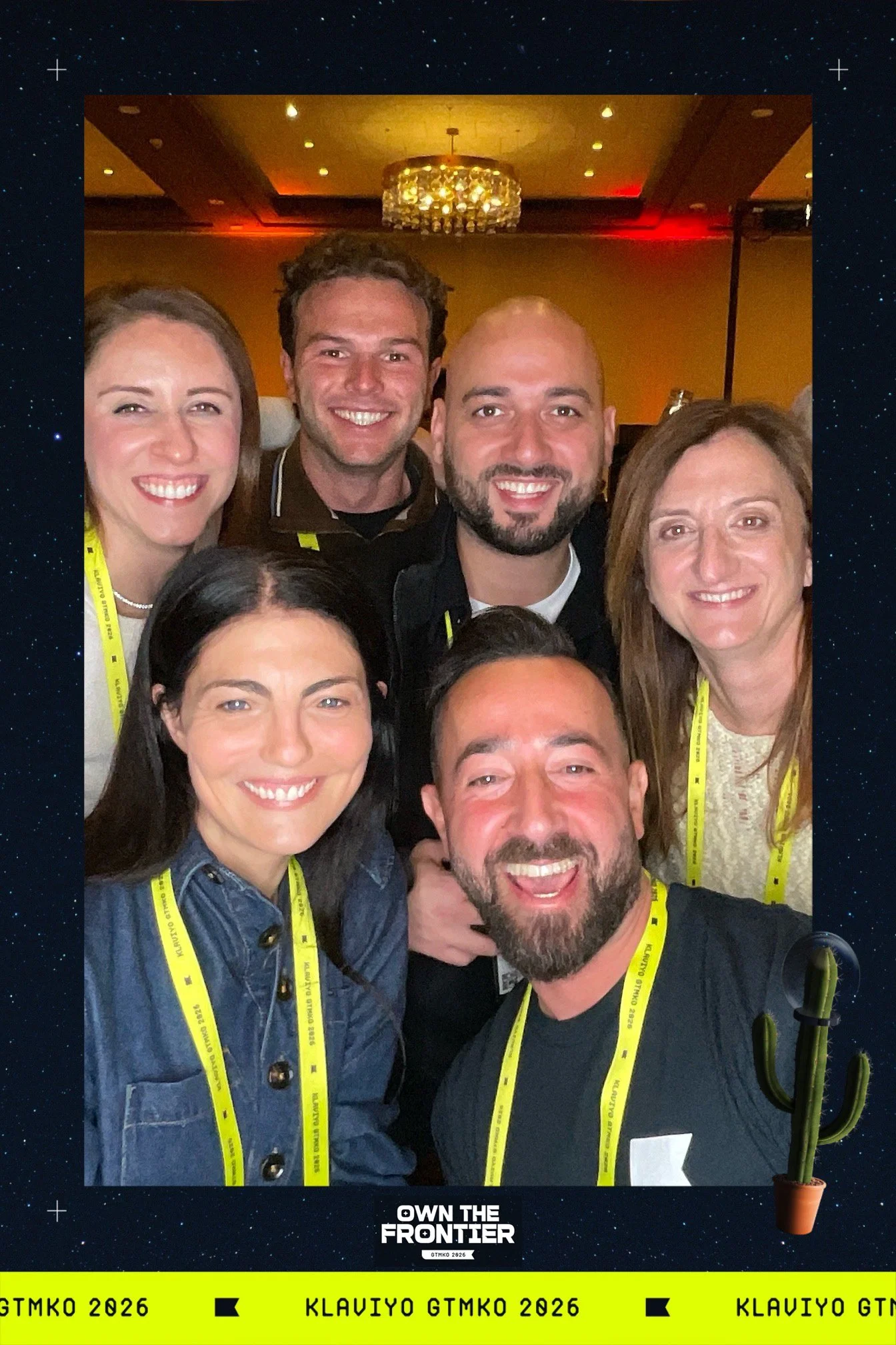 A group of seven smiling people taking a selfie at an event, with a decorative chandelier in the background and yellow lanyards around their necks that read "Own the Frontier" and "Klaviyo GTMK 2026".