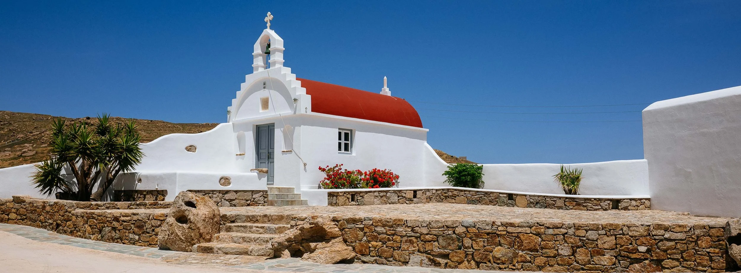 White church with red domed roof and cross on top, set against a clear blue sky, with stone steps and decorative plants in front.