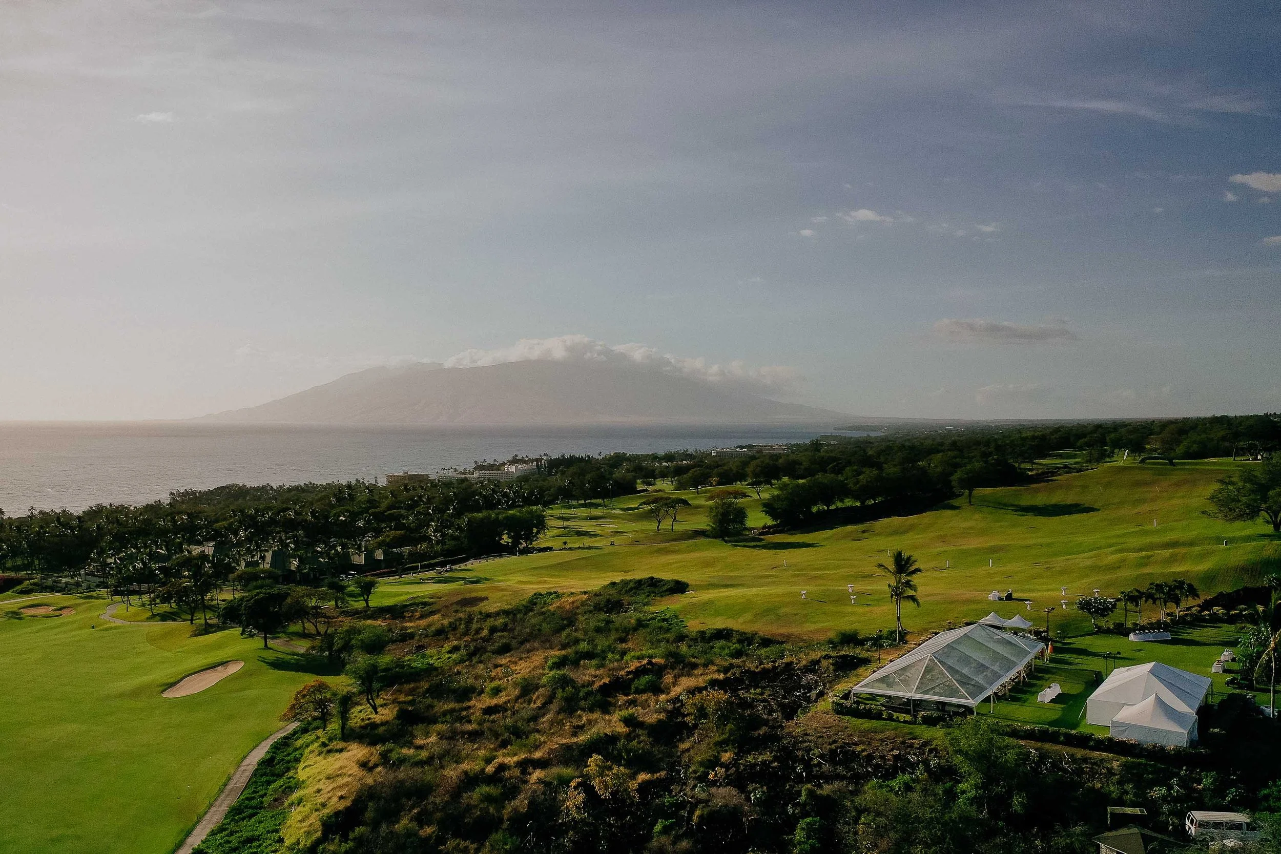 Scenic view of a golf course near the coast with a mountain in the background, green fairways, trees, and a few tents or structures in the foreground under a partly cloudy sky.